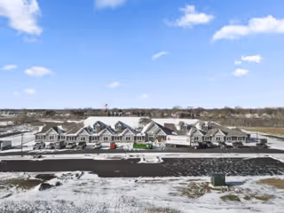 Wide exterior view of a large senior living facility building with multiple peaked roofs, surrounded by a snowy landscape under a partly cloudy blue sky.