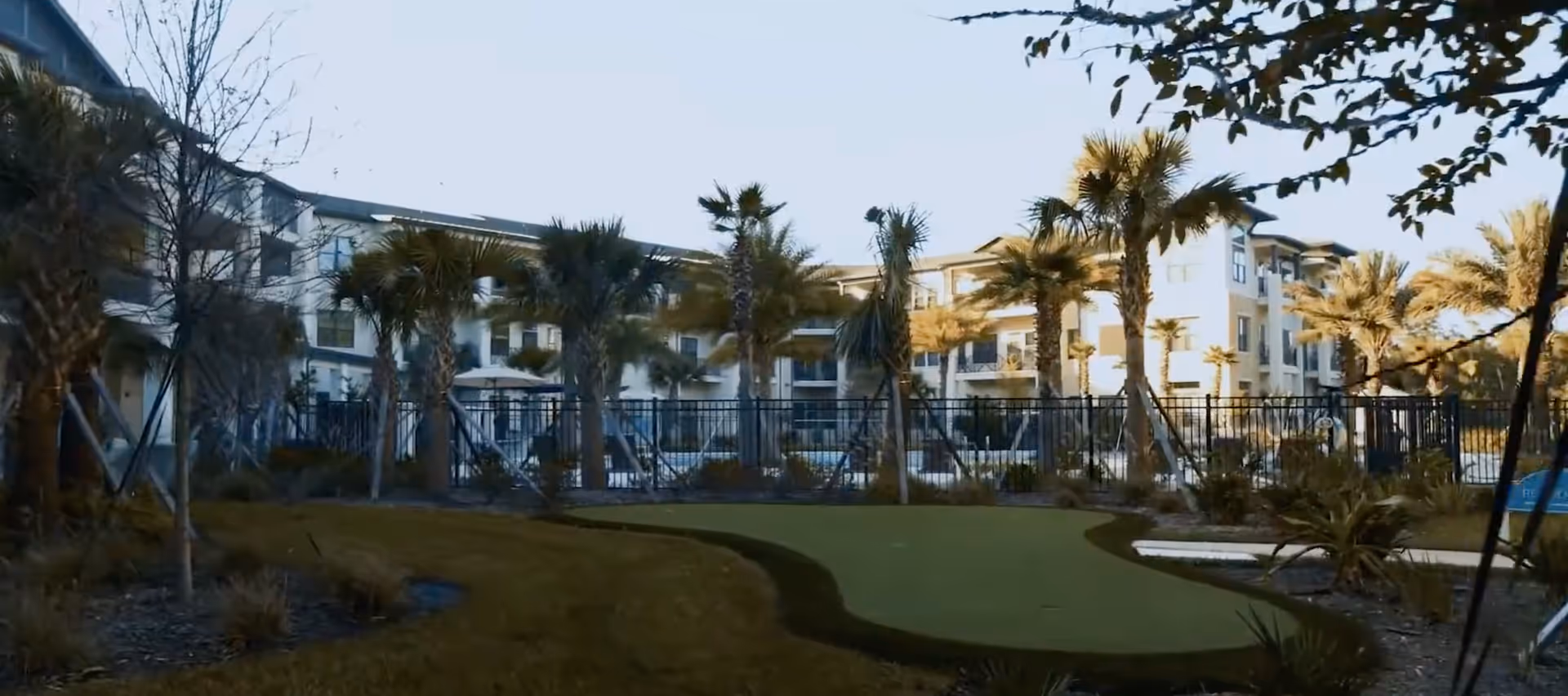 Outdoor view of a senior living facility with a putting green in the foreground, palm trees, and a fenced pool area surrounded by a multi-story building under clear sky.