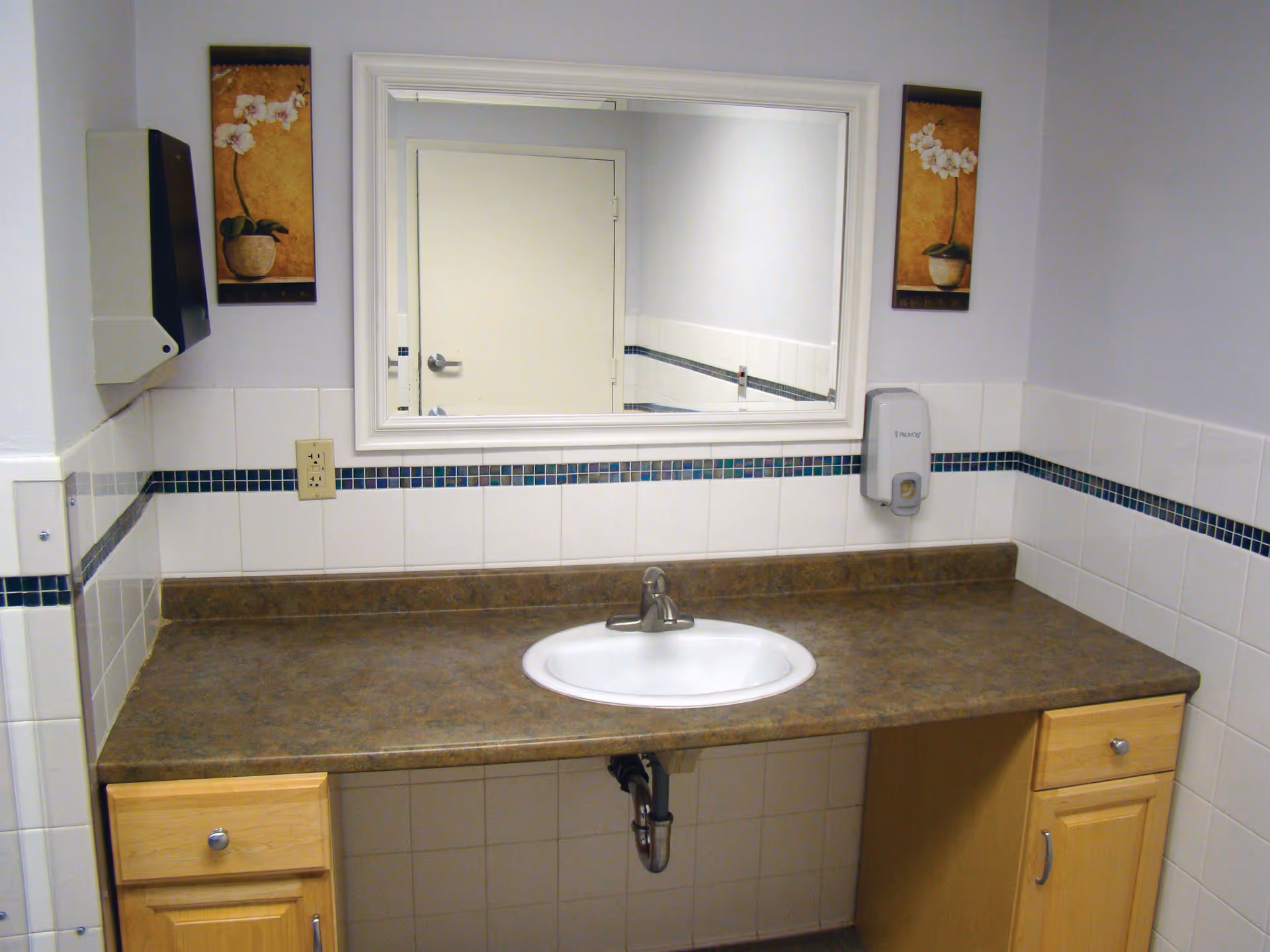 A clean bathroom sink area with a brown countertop, a white oval sink, and wooden cabinets underneath. Above the sink is a large rectangular mirror reflecting a closed door. The walls are tiled with white tiles and a decorative blue tile border. On the left wall is a paper towel dispenser, and on the right wall is a soap dispenser. Two framed pictures with floral designs hang on either side of the mirror.