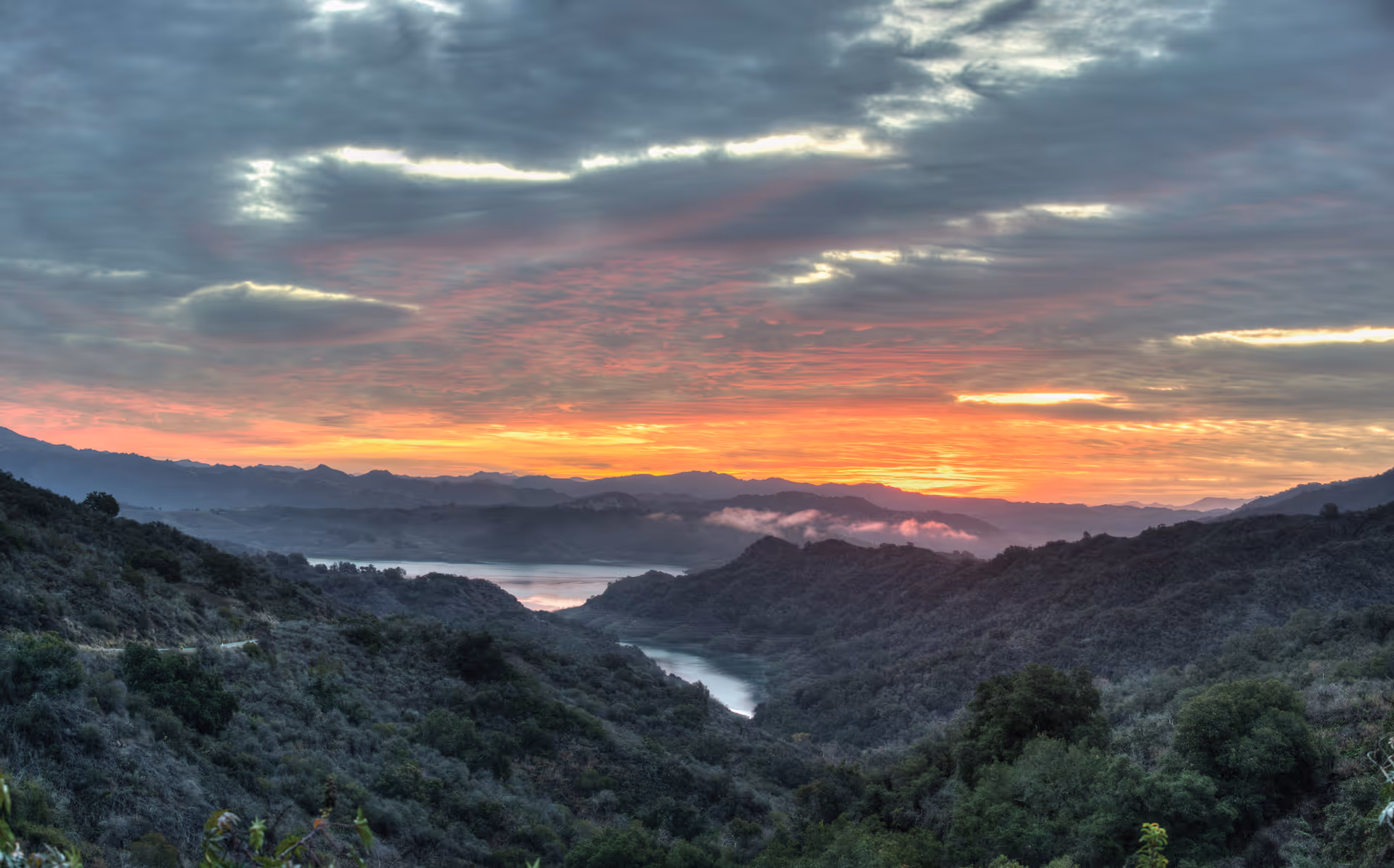 Sunset over rolling green hills and a winding reservoir valley beneath dramatic layered clouds.