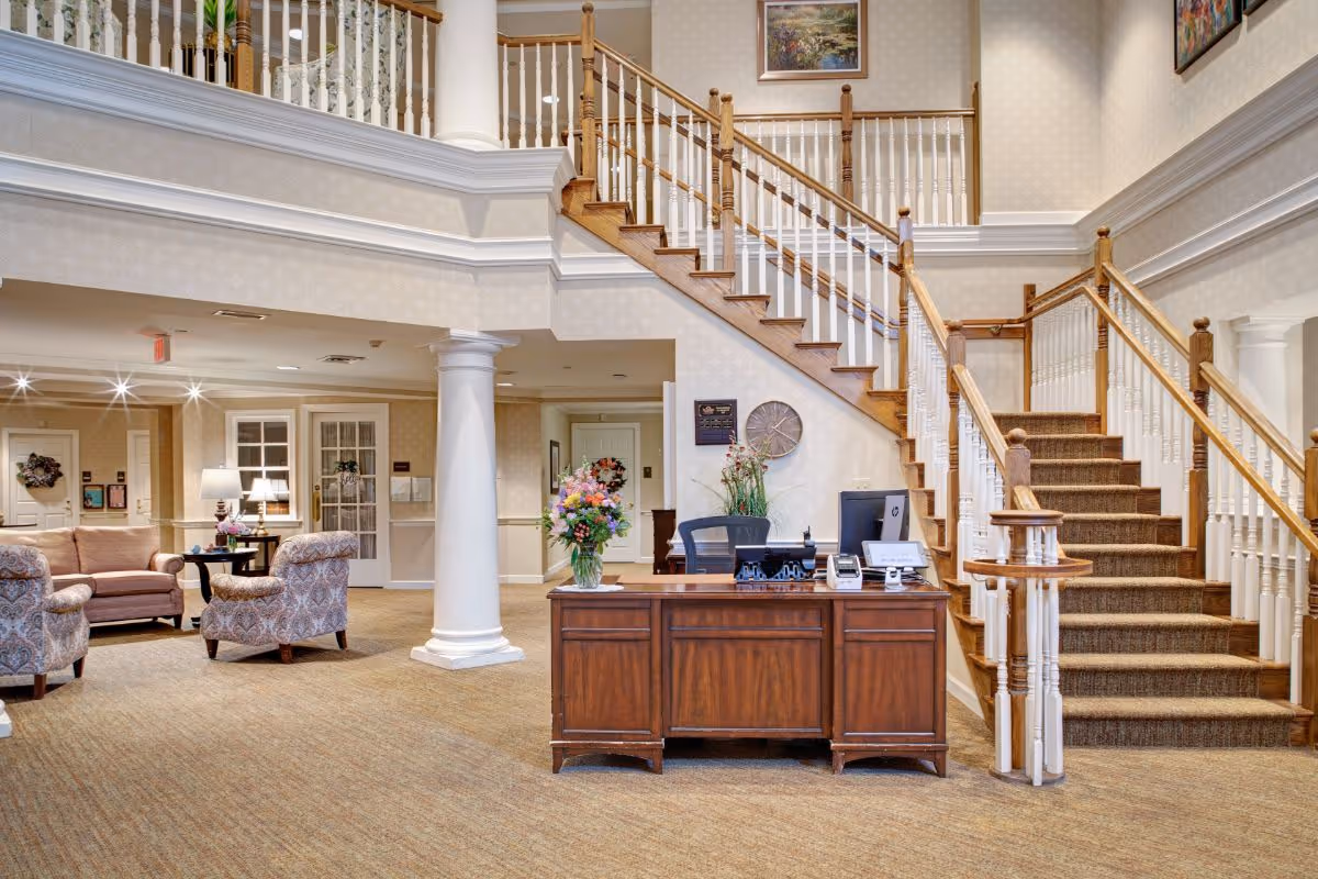 Interior view of a senior living facility lobby with a wooden reception desk in front of a staircase with wooden handrails and white balusters. To the left, there is a seating area with upholstered chairs and a sofa, a side table with a lamp, and decorative wreaths on the walls. The space is well-lit with ceiling lights and decorated with floral arrangements and framed artwork.