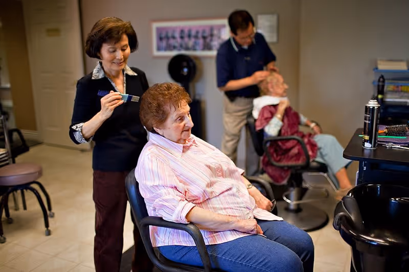 Two elderly women seated in salon chairs getting their hair styled by two hairstylists in a salon setting with hair care products visible on a counter.