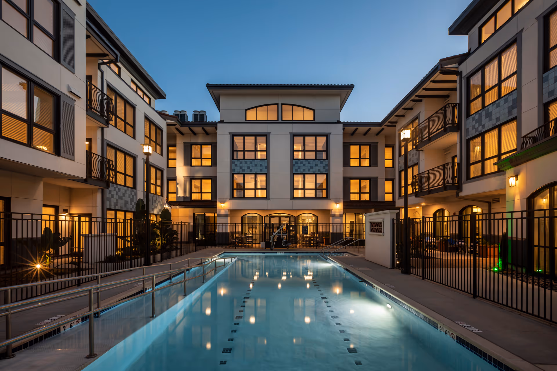 Evening view of a modern senior living facility courtyard with a swimming pool in the center, surrounded by a black metal fence and multi-story buildings with large windows illuminated from inside.