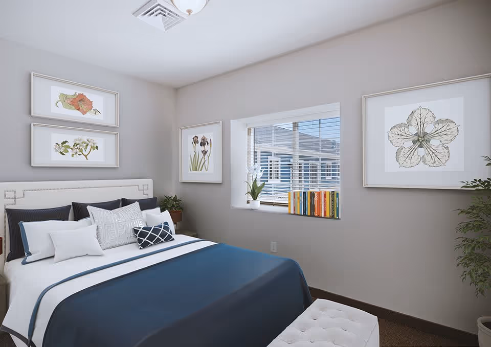 A neatly made bed with white and dark blue bedding in a bedroom. The walls are light gray and decorated with framed botanical prints. A window with blinds lets in natural light, and a small plant and a row of books are placed on the windowsill. A white cushioned bench is at the foot of the bed, and there is a potted plant in the corner.