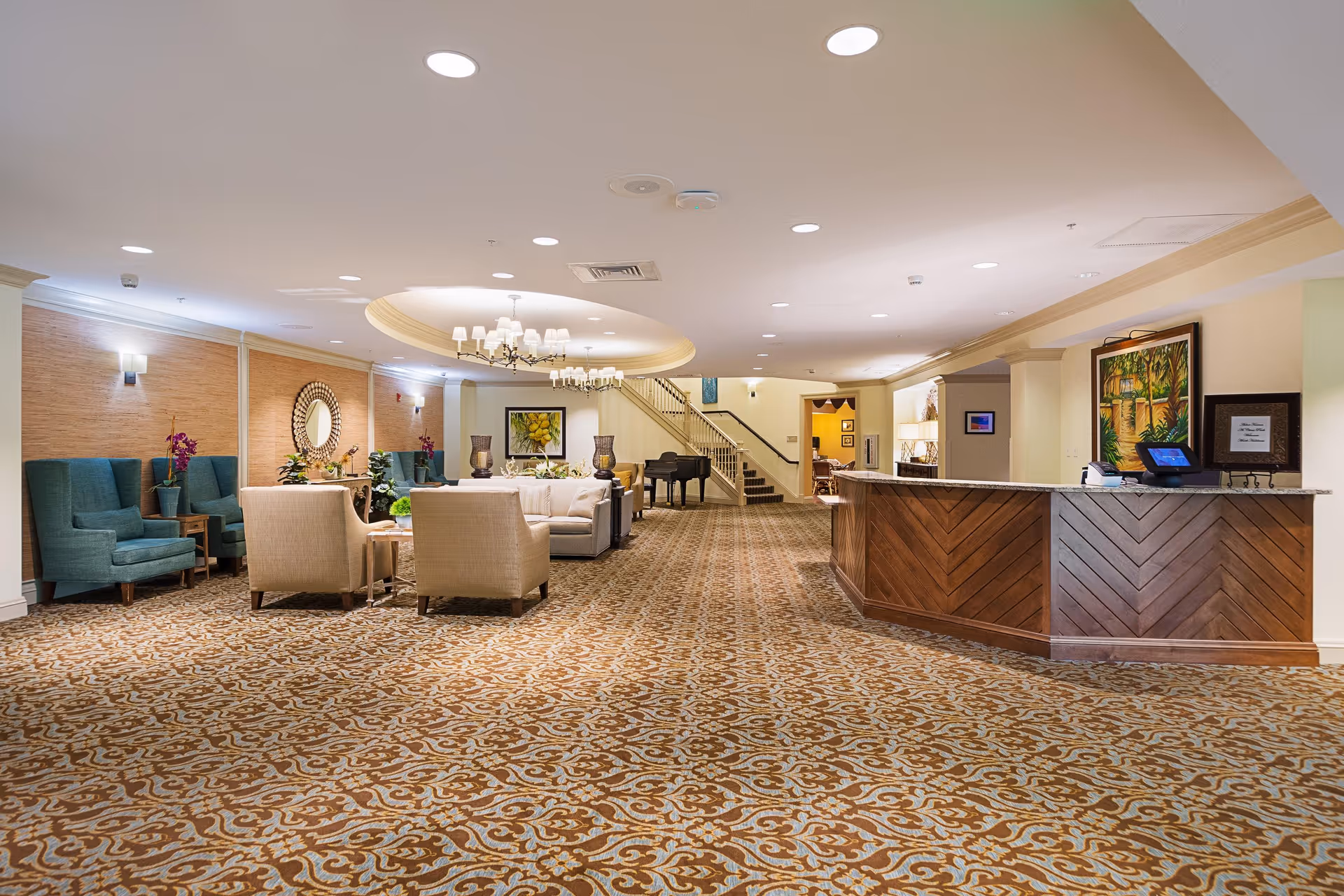 Spacious senior living facility lobby with patterned carpet, a wooden reception desk on the right, several seating areas with beige and teal armchairs, a chandelier hanging from the ceiling, decorative plants, and a grand piano near a staircase in the background.
