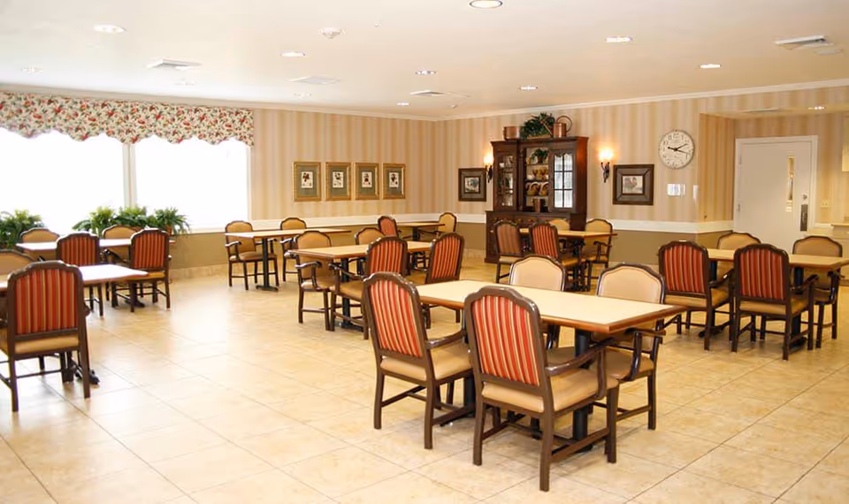 A bright dining room with multiple tables and chairs arranged neatly. The room has large windows with floral valances, beige tiled floors, striped wallpaper, framed pictures on the walls, a clock, and a wooden cabinet with decorative items.