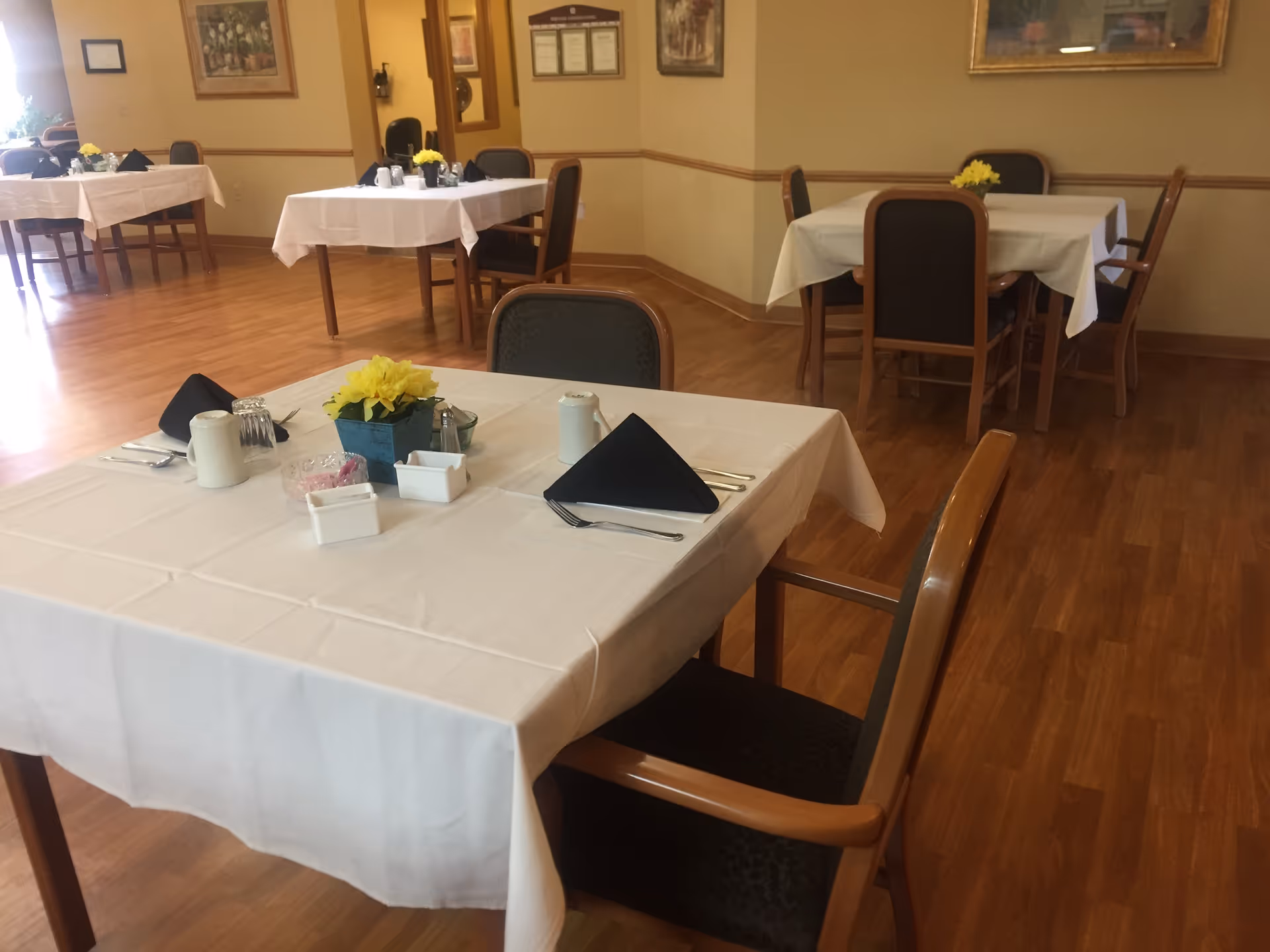 A dining room with several tables covered in white tablecloths, each set with black folded napkins, silverware, and small flower arrangements. The room has wooden flooring, framed artwork on the walls, and chairs with wooden arms and dark upholstery.