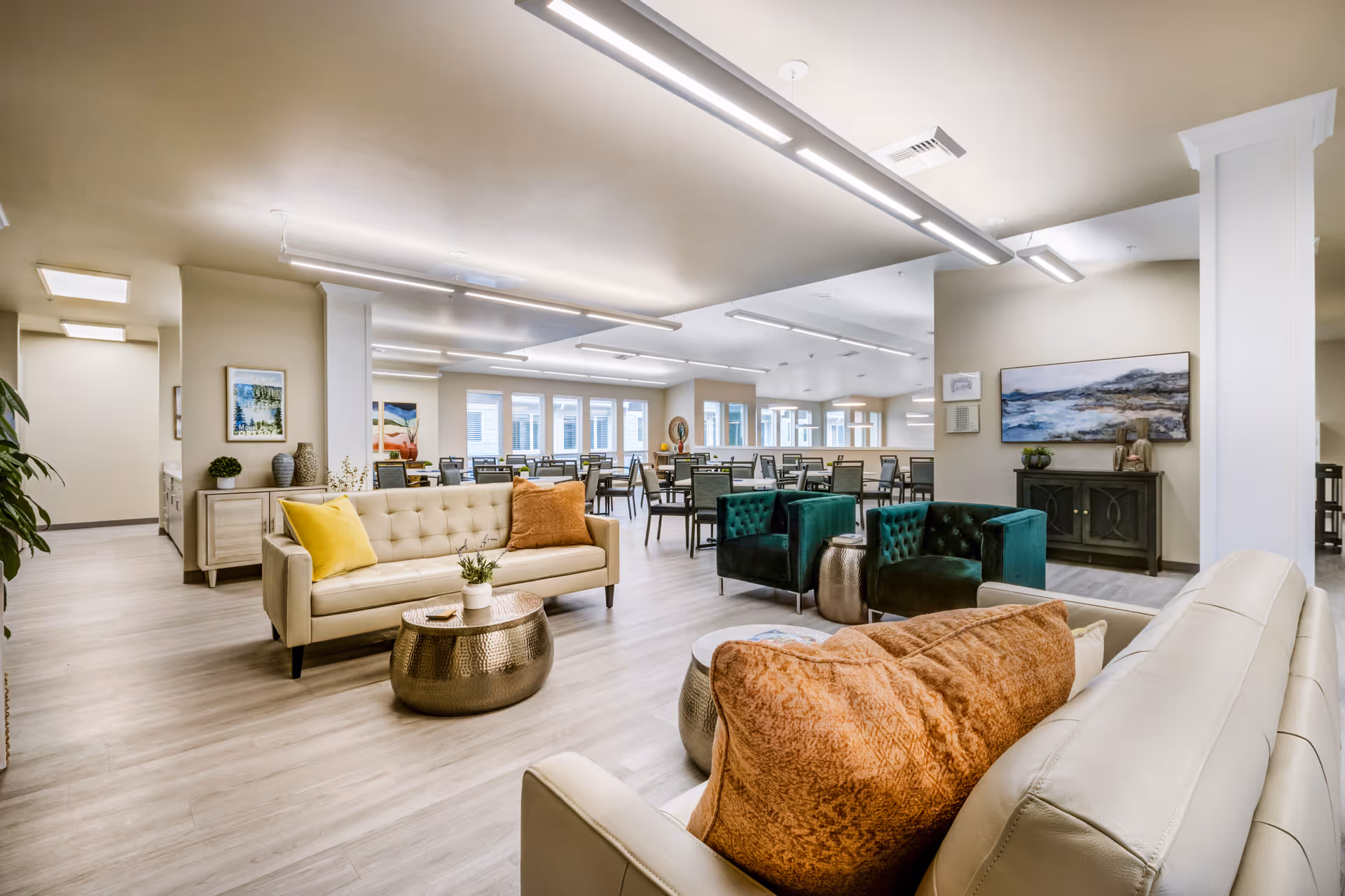 A spacious and well-lit senior living common area featuring beige leather sofas with orange and yellow pillows, two green velvet armchairs, and round metallic coffee tables. In the background, there are multiple dining tables and chairs arranged neatly near large windows, with modern ceiling lights illuminating the space. The walls are decorated with framed artwork and plants, creating a welcoming and comfortable atmosphere.