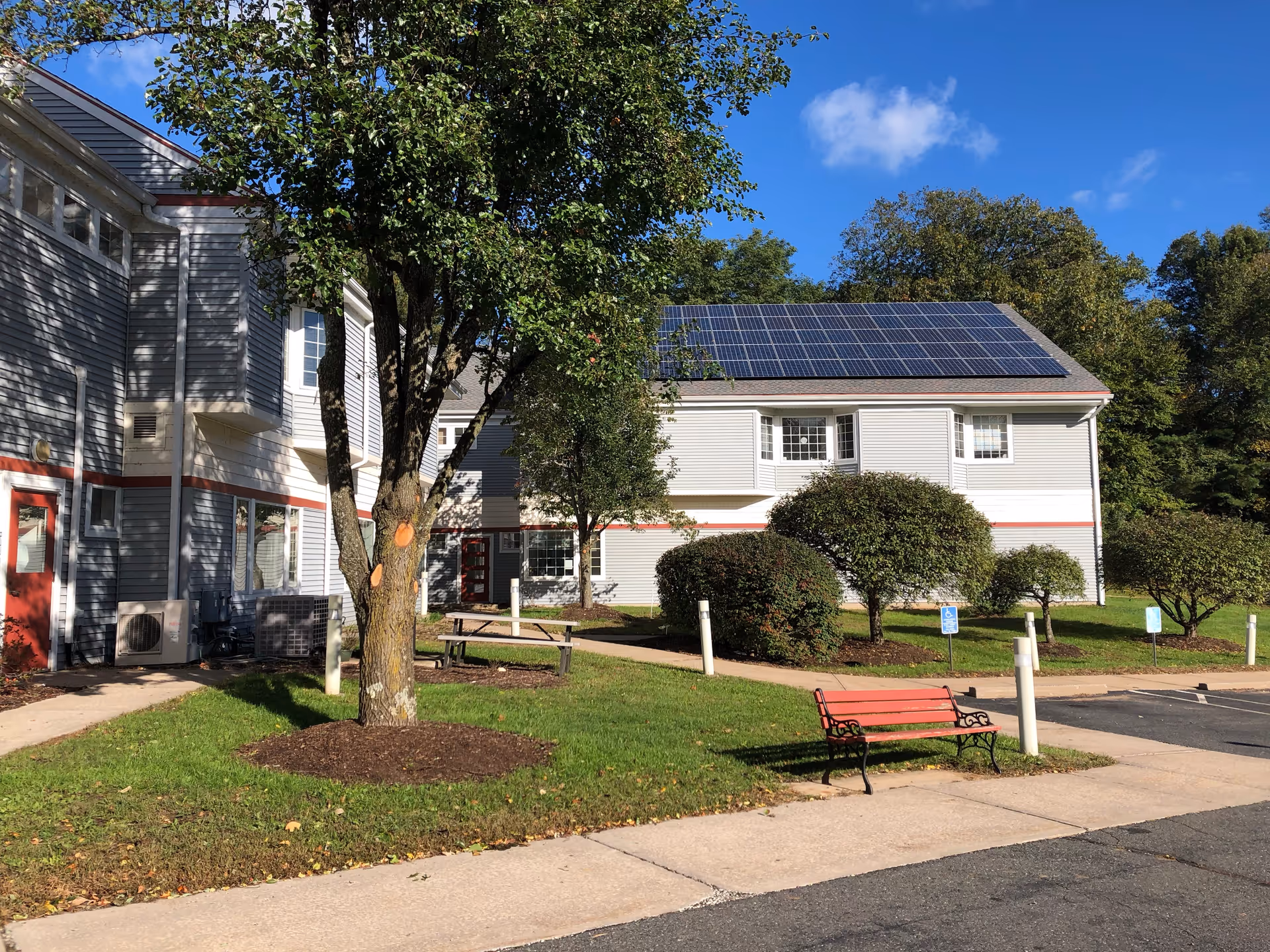 Exterior view of a senior living facility building with gray siding and red trim. The building has solar panels installed on the roof. There is a tree and several bushes in the grassy area in front of the building, along with a red bench and a picnic table. The sky is clear and blue.