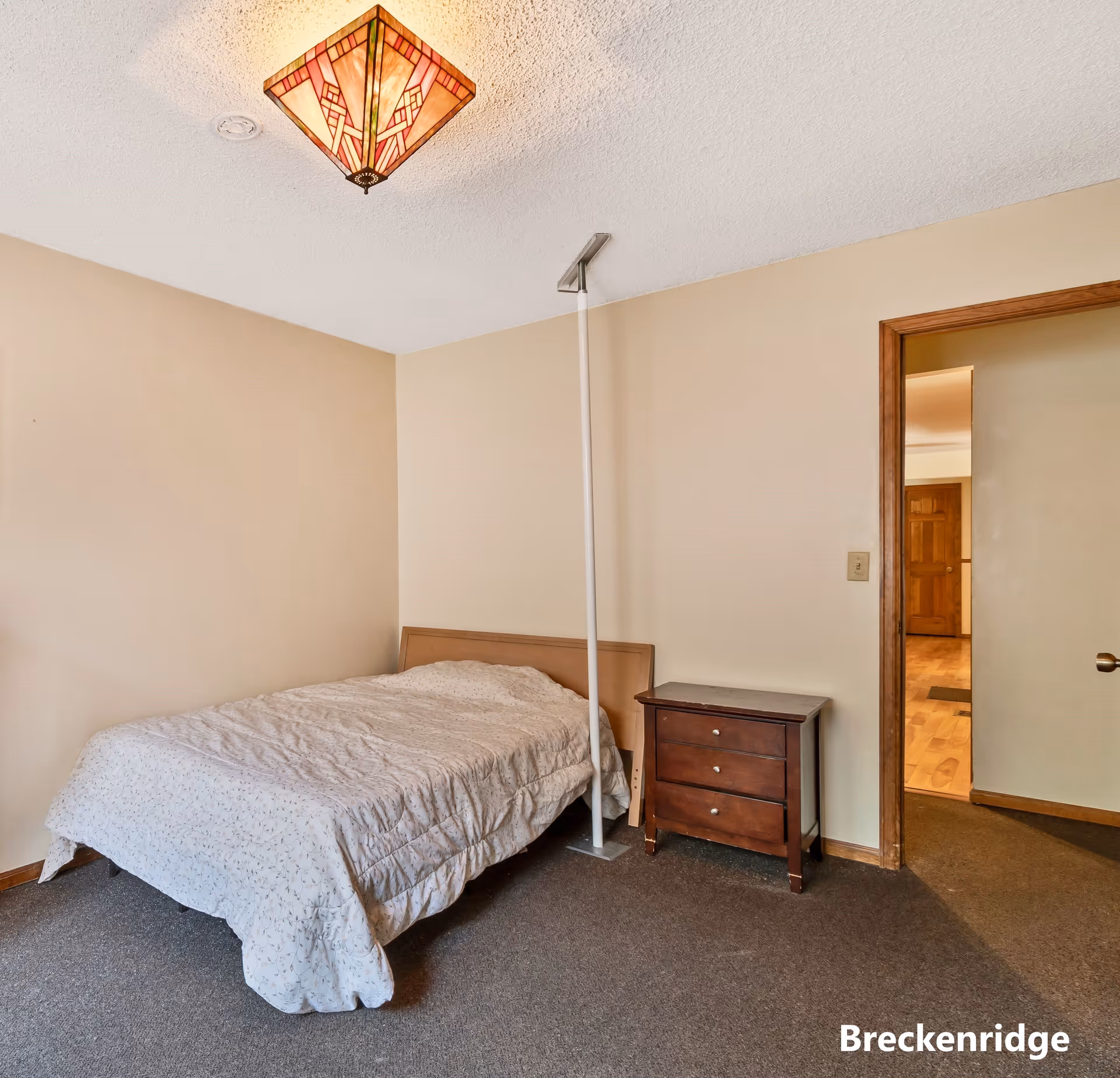 A simple bedroom with a bed covered in a light-colored quilt, a wooden headboard, a dark wooden nightstand with three drawers, beige walls, a carpeted floor, and a decorative ceiling light fixture. There is a white support pole next to the bed and an open doorway leading to another room with wooden flooring.