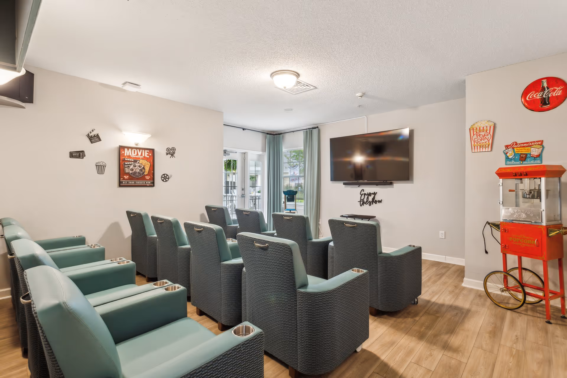 Small screening room with rows of teal recliner chairs facing a wall-mounted TV, movie-themed decor, and a popcorn machine.
