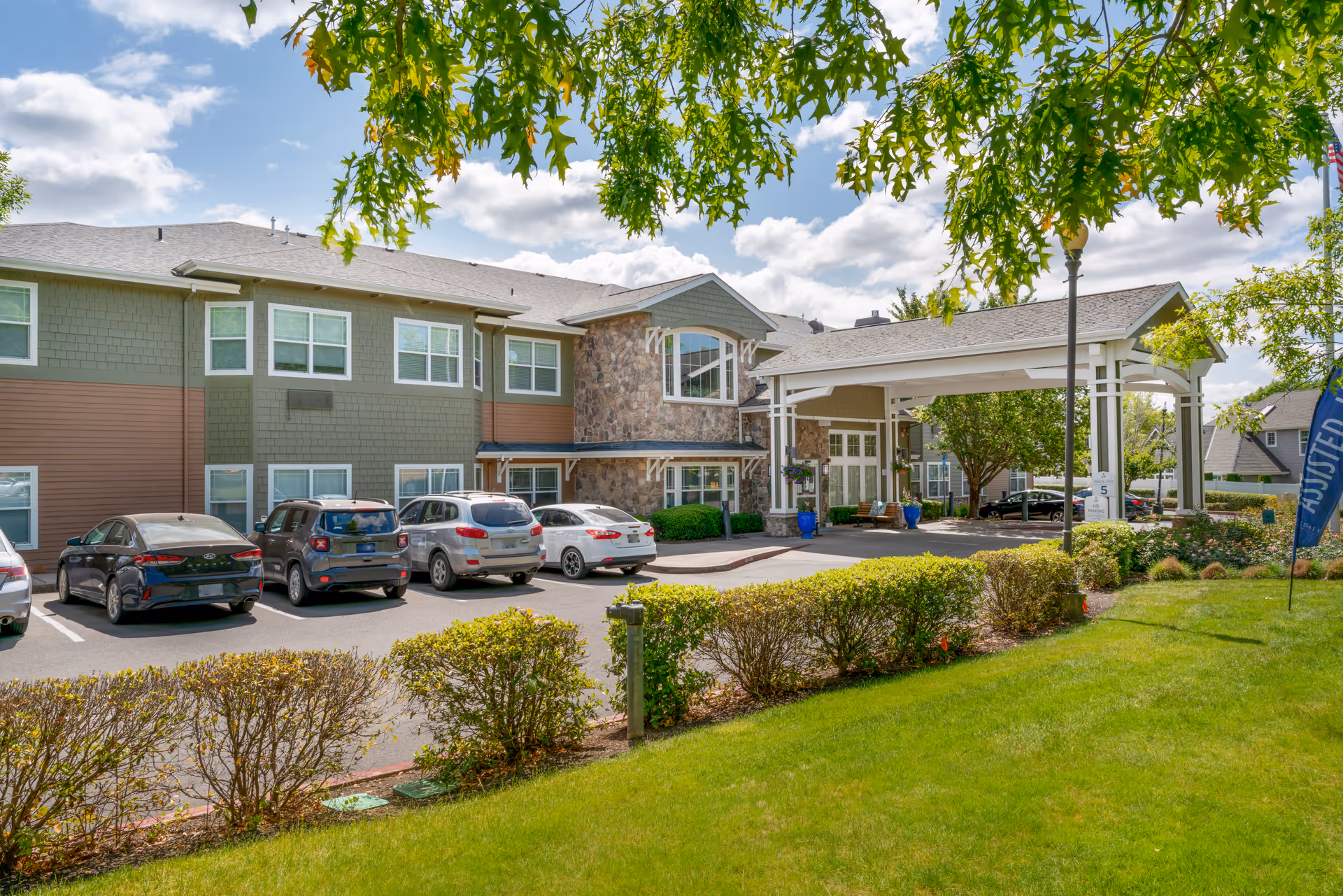 Exterior view of Brookdale Newberg senior living facility showing a two-story building with green and brown siding, stone accents, and multiple windows. Several cars are parked in front of the building under a partly cloudy sky. Green bushes and a well-maintained lawn are visible in the foreground, with tree branches hanging down from the top of the image.