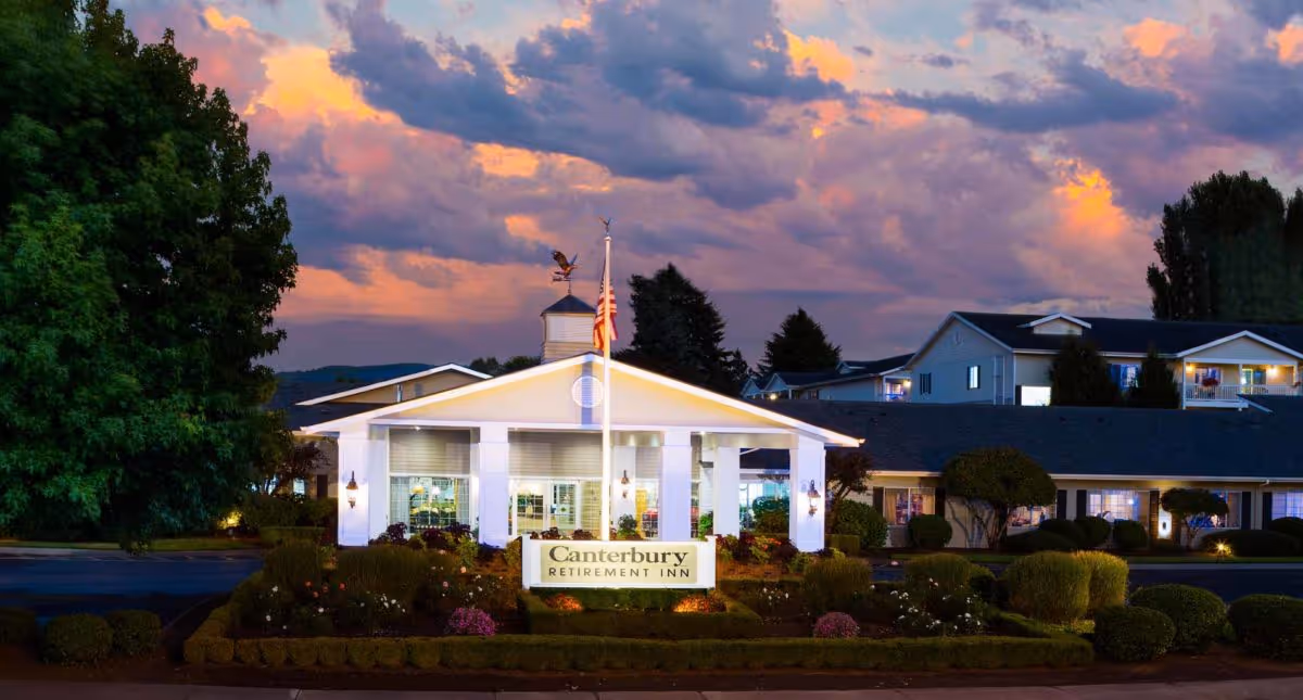 Exterior view of Canterbury Retirement Inn at dusk with a dramatic cloudy sky. The building is white with a peaked roof and an American flag on a pole in front. Well-maintained bushes and flowers surround the entrance.