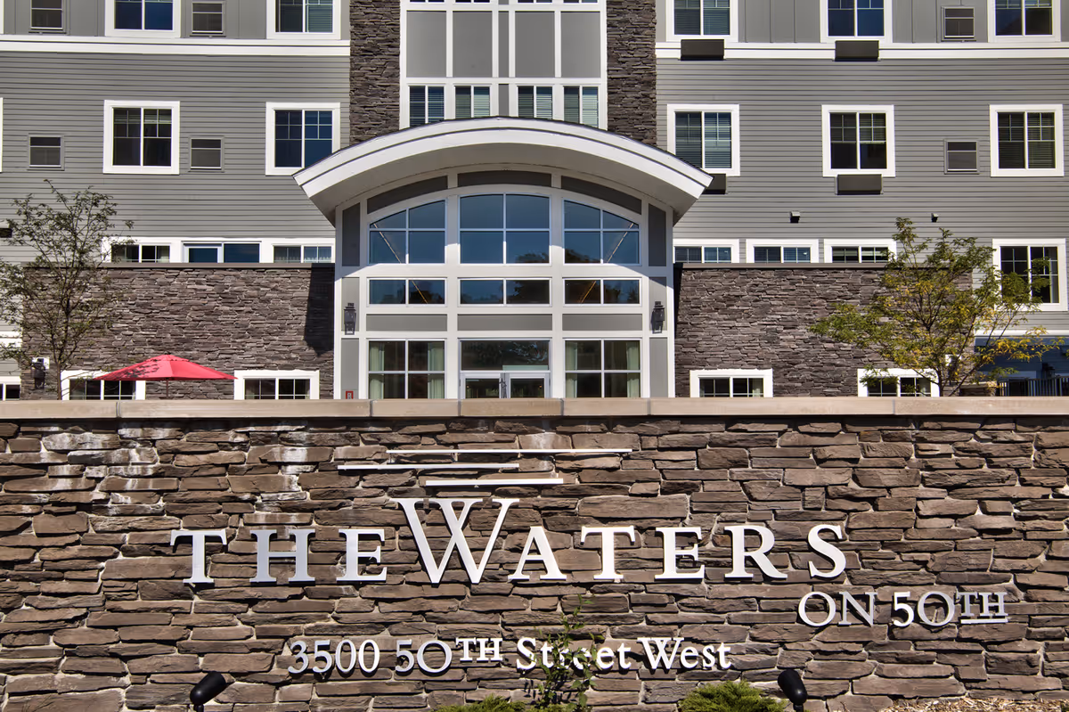 Exterior view of The Waters on 50th senior living facility showing a stone wall with the facility name and address, and a multi-story building with large windows and an arched entrance in the background.