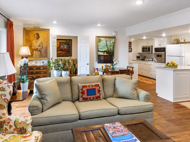 Open, well-lit living room with a green sofa and coffee table facing an adjoining kitchen and dining area with framed paintings and plants.