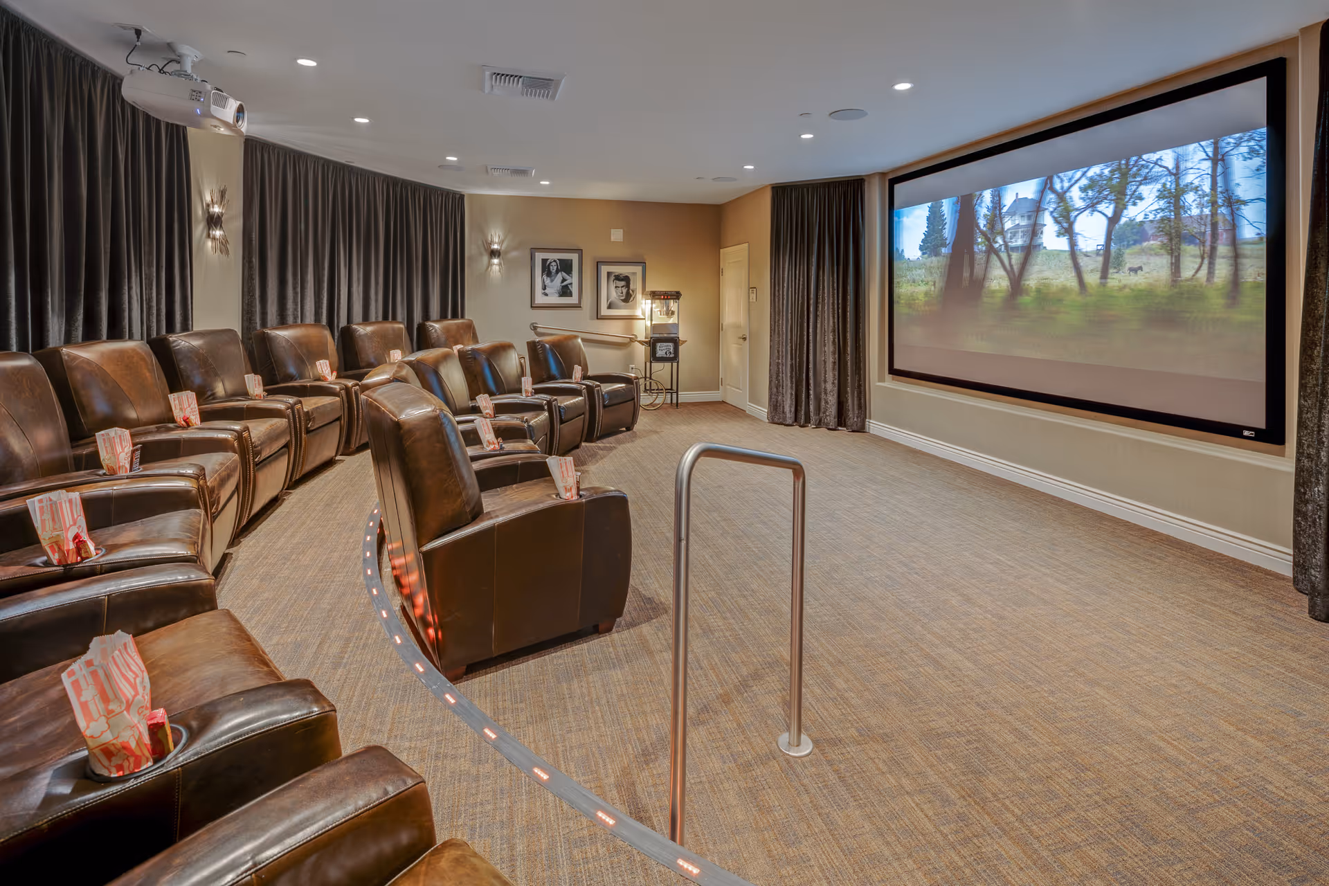 A cozy movie theater room with two rows of brown leather recliner chairs, each with a popcorn bag placed on the armrest cup holder. The room has a large projector screen displaying an outdoor scene with trees and a house. There is a popcorn machine in the back corner, framed black and white photos on the wall, and dark curtains covering the windows.