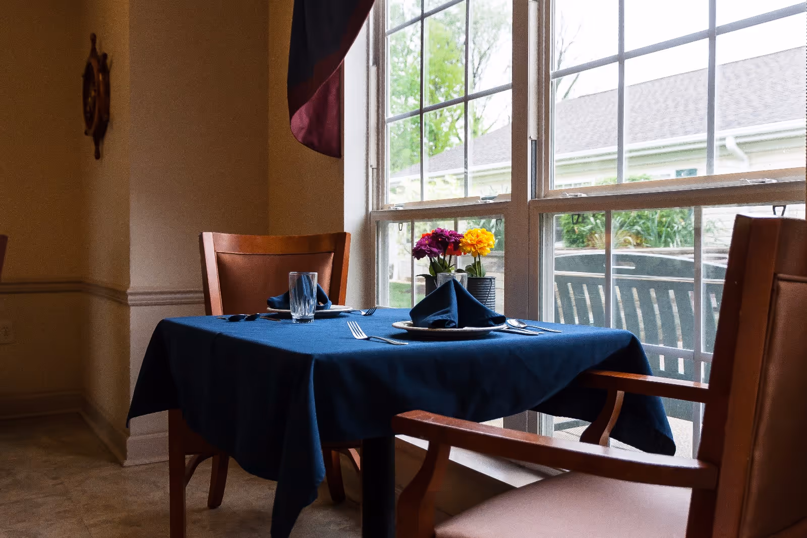 A dining table set for two with a dark blue tablecloth, folded napkins on plates, silverware, and a glass. There is a small vase with colorful flowers on the table. The table is next to large windows showing an outdoor patio area with chairs and greenery.