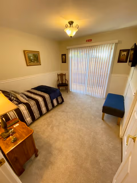 A small, neatly arranged bedroom with a single bed covered in a striped blanket and a dark blue throw. There is a wooden nightstand with a lamp and some small items on it next to the bed. A wooden chair is placed against the wall near a sliding glass door with vertical blinds. A blue cushioned bench is positioned near the door. The walls are light-colored with white wainscoting, and there are framed pictures hanging on the walls. The room is carpeted and well-lit by a ceiling light fixture.