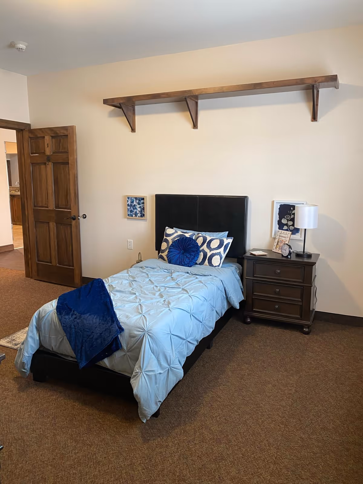 A bedroom with a single bed featuring a light blue comforter and decorative pillows in blue and white patterns. Next to the bed is a dark wooden nightstand with a lamp, a clock, and framed pictures. The room has beige walls, brown carpet, a wooden door slightly open, and a long wooden shelf mounted high on the wall above the bed.