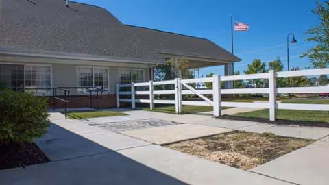 Single-story facility exterior with white fence, sidewalk, and American flag on a sunny day.