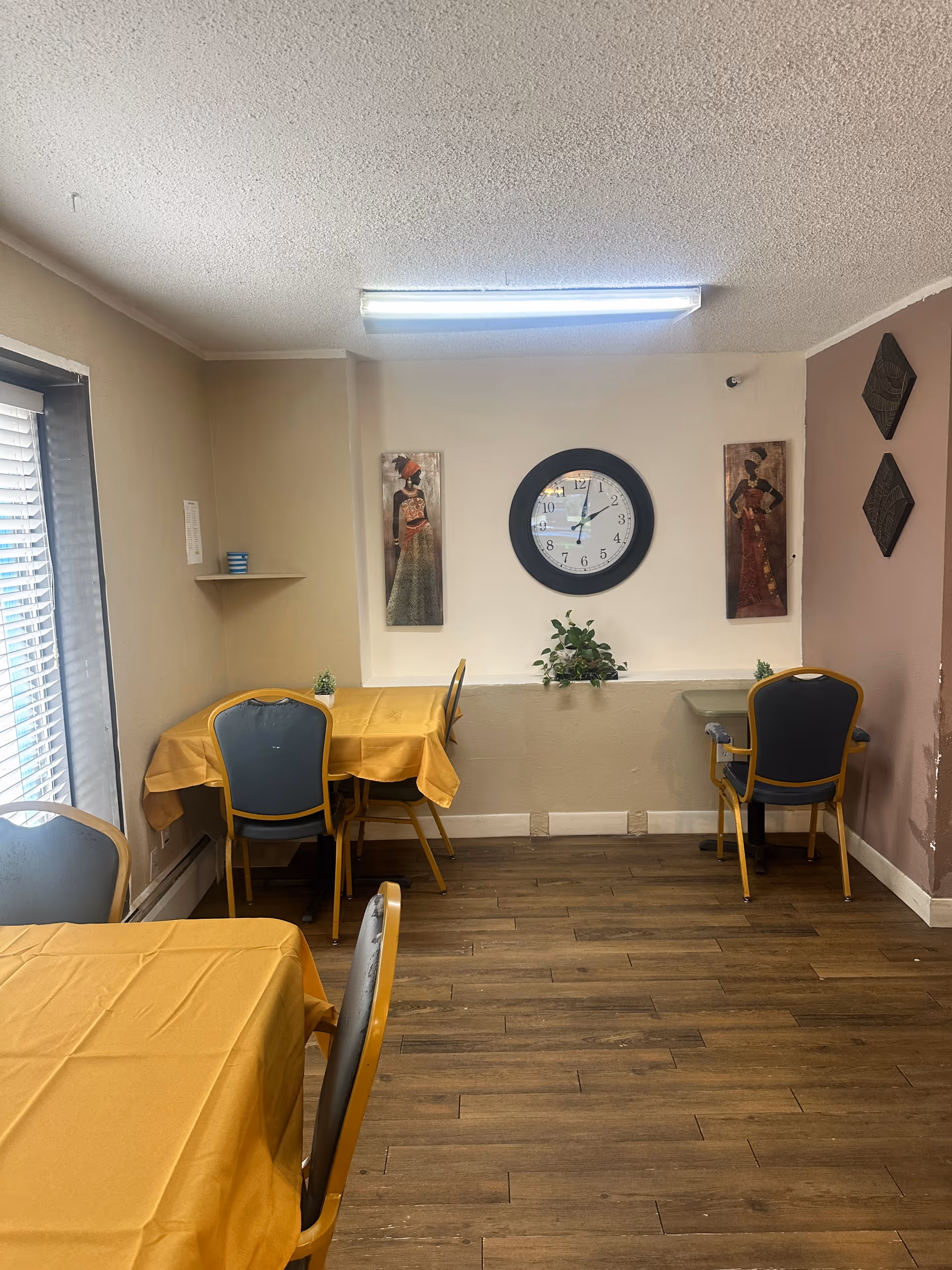 A small dining area with three tables covered in yellow tablecloths and blue chairs. The room has wooden flooring and beige walls with decorative artwork and a round wall clock showing the time as 12:05. There is a small plant on a ledge beneath the clock and a window with blinds on the left side.