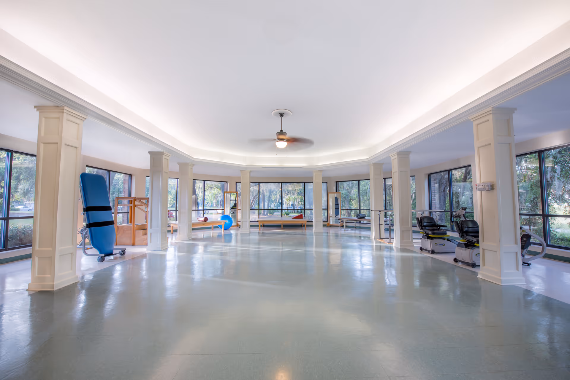 A spacious, well-lit rehabilitation or physical therapy room with large windows, white columns, and exercise equipment including recumbent bikes, therapy tables, and balance tools. The room has a ceiling fan and a light-colored floor.
