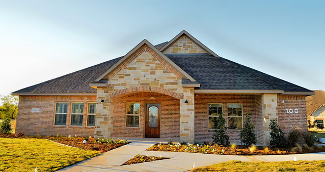 Front exterior view of a single-story brick and stone building with a peaked roof, a wooden front door with glass panels, and multiple windows. The building is surrounded by a landscaped lawn with small bushes and flowers along the walkway leading to the entrance.