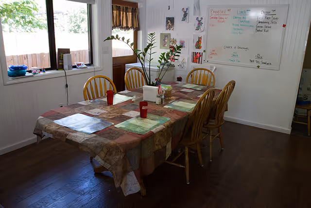Dining room with a rectangular table covered by a patchwork tablecloth, surrounded by eight wooden chairs. The table has placemats, red cups, a napkin holder, and a small plant. Large windows with brown valances let in natural light. A whiteboard on the wall displays a menu and some artwork.