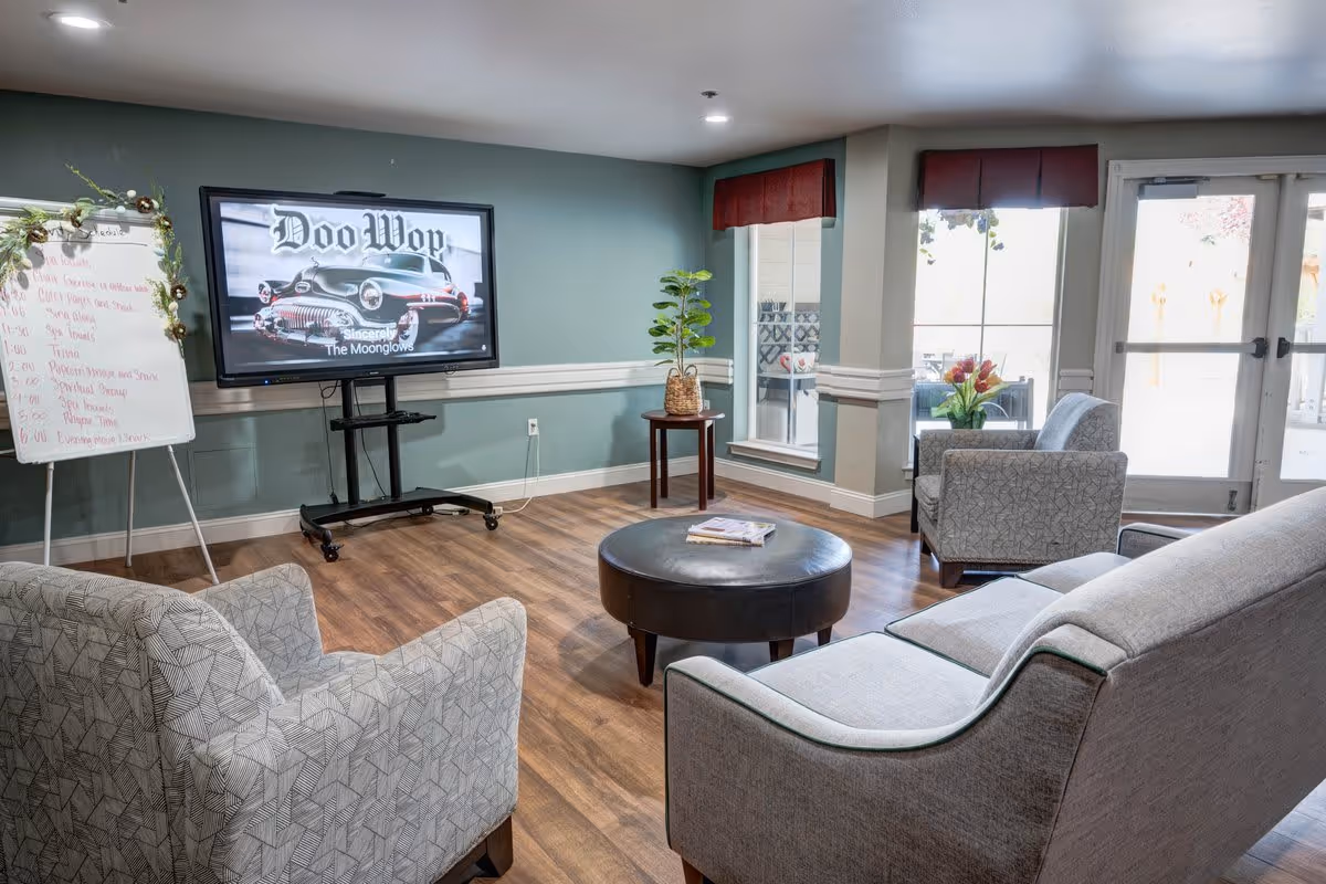 A cozy living room area with a gray patterned armchair, a gray sofa, and a round black ottoman in the center. A flat-screen TV on a stand displays an image with the text 'Doo Wop Sincerely The Moonglows.' There is a whiteboard with a schedule written on it to the left of the TV. The room has wood flooring, green walls with white trim, two windows with red valances, and a glass door leading outside. A small table with a potted plant is placed between the windows.