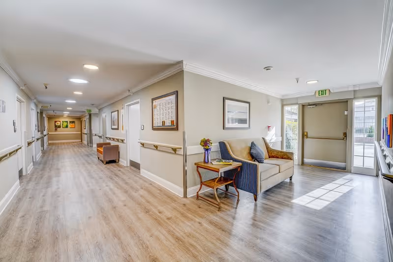 A bright and clean hallway in a senior living facility with light wood flooring and beige walls. There is a small seating area with a beige and blue loveseat, a side table with a vase of flowers, and framed artwork on the walls. The hallway has handrails on both sides and leads to an exit door with windows letting in natural light.