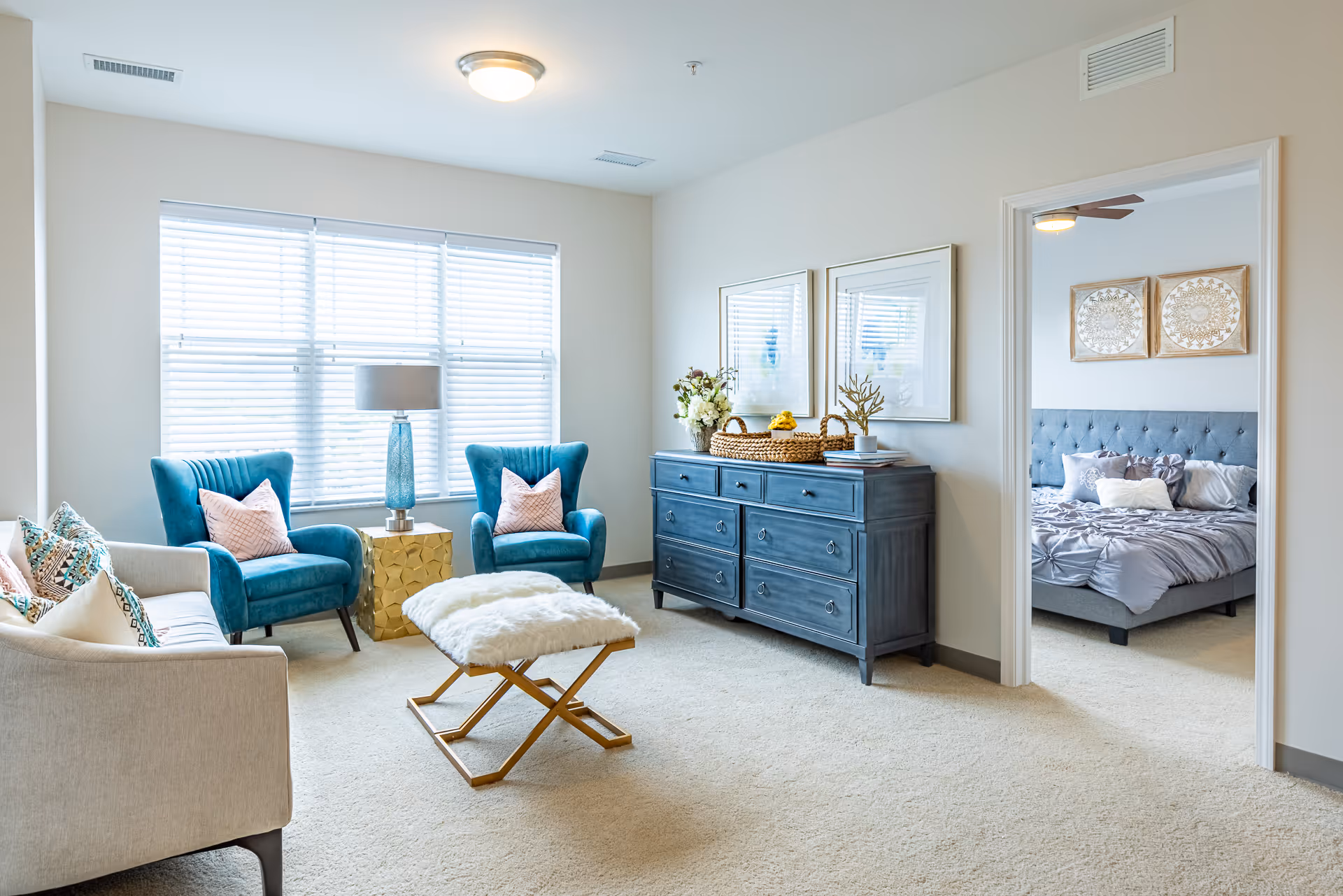 A bright living room with a beige carpet, two teal armchairs with pink pillows, a beige sofa with patterned pillows, a gold side table with a blue lamp, and a furry white ottoman. A blue dresser with decorative items and two framed pictures hangs on the wall. Through an open doorway, a bedroom with a gray tufted bed and two decorative wall hangings is visible.