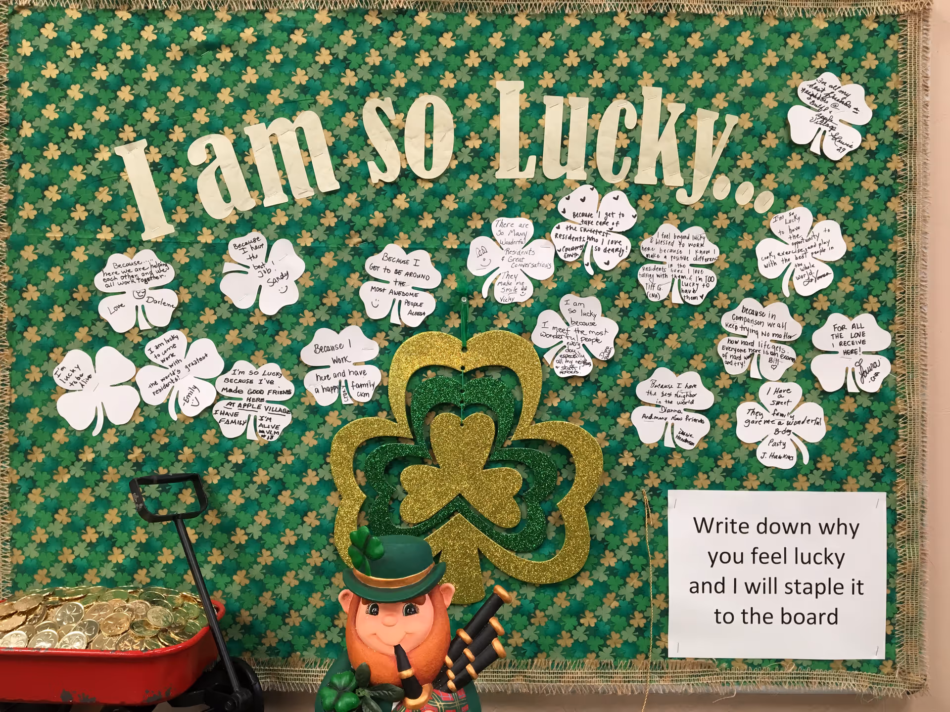 St. Patrick's Day bulletin board reading 'I am so Lucky!' decorated with paper shamrocks of handwritten notes, a glitter clover centerpiece, a leprechaun figure and a sign inviting residents to write why they feel lucky.