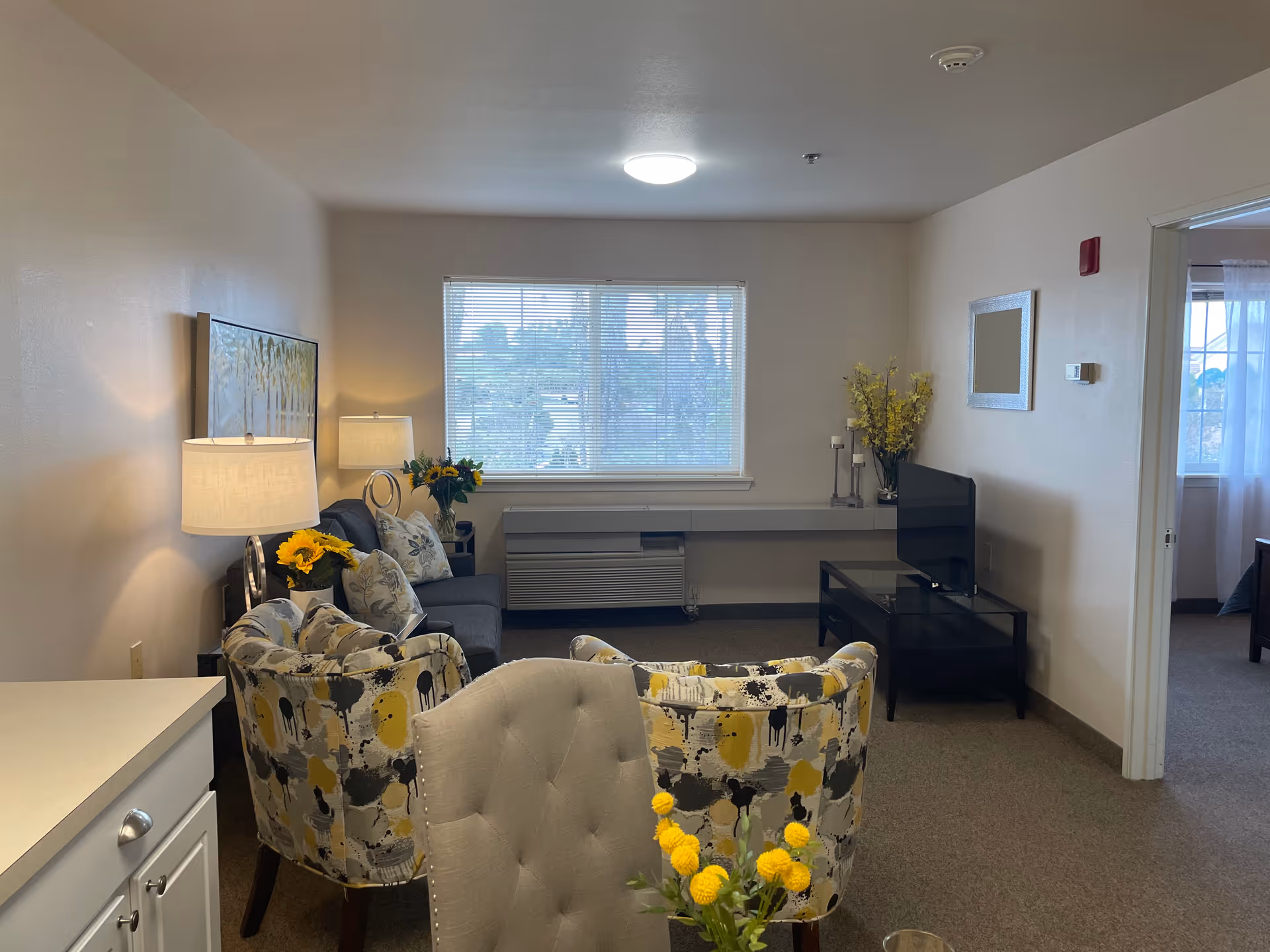 A cozy living room with a window letting in natural light. The room features a gray sofa, two patterned armchairs with yellow and gray accents, a small black TV stand with a flat-screen TV, a lamp on a side table, and decorative flowers. A doorway on the right leads to a bedroom with a bed visible.