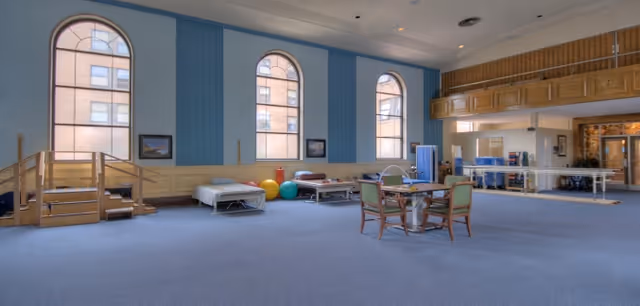 A spacious rehabilitation or therapy room with large arched windows letting in natural light. The room features therapy beds, exercise balls, parallel bars for walking practice, a small table with chairs, and wooden stairs for physical therapy exercises. The walls are painted blue with wood paneling, and the carpet is blue.