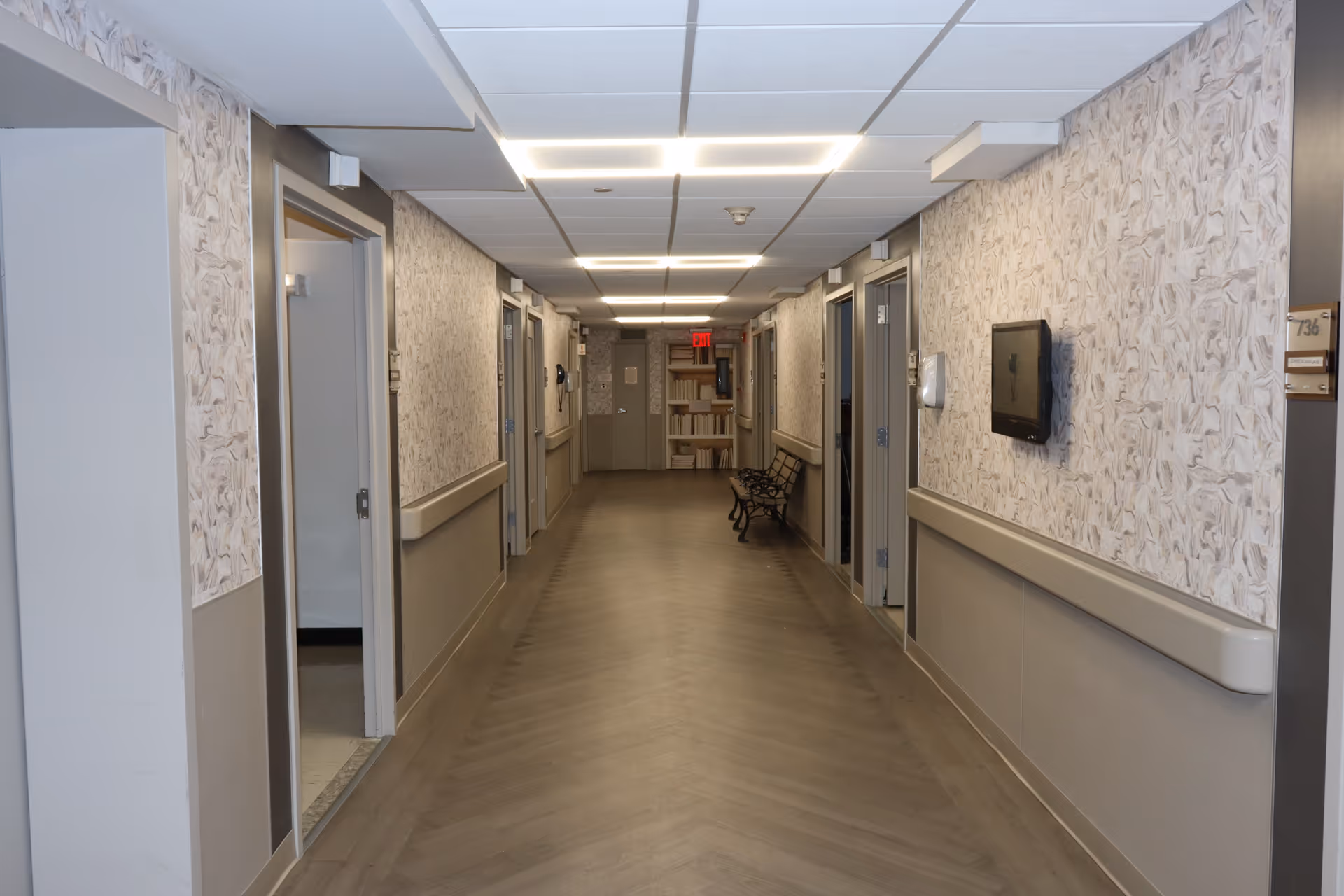A long, well-lit hallway in a rehabilitation and nursing facility with patterned wallpaper, handrails on both sides, several open doors leading to rooms, a wall-mounted TV, and a bench near the end of the corridor.