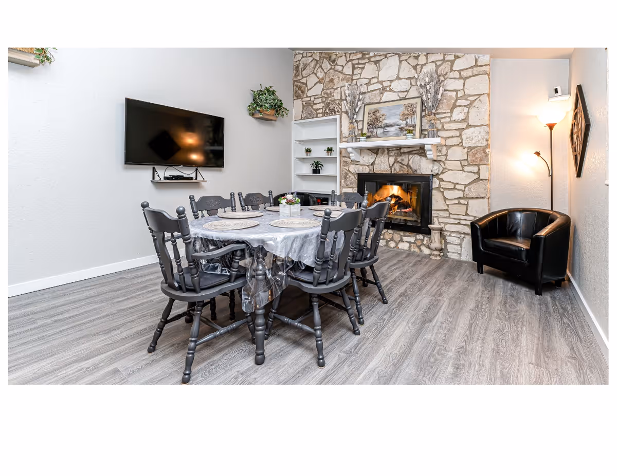A cozy dining area with a round table covered with a plastic tablecloth and six black wooden chairs around it. The room features a stone fireplace with a fire burning, a painting above the mantel, and decorative plants. A black leather armchair is placed near a floor lamp in the corner. A flat-screen TV is mounted on the wall opposite the fireplace, and there are shelves with small plants next to the fireplace. The floor has a wood-like finish.