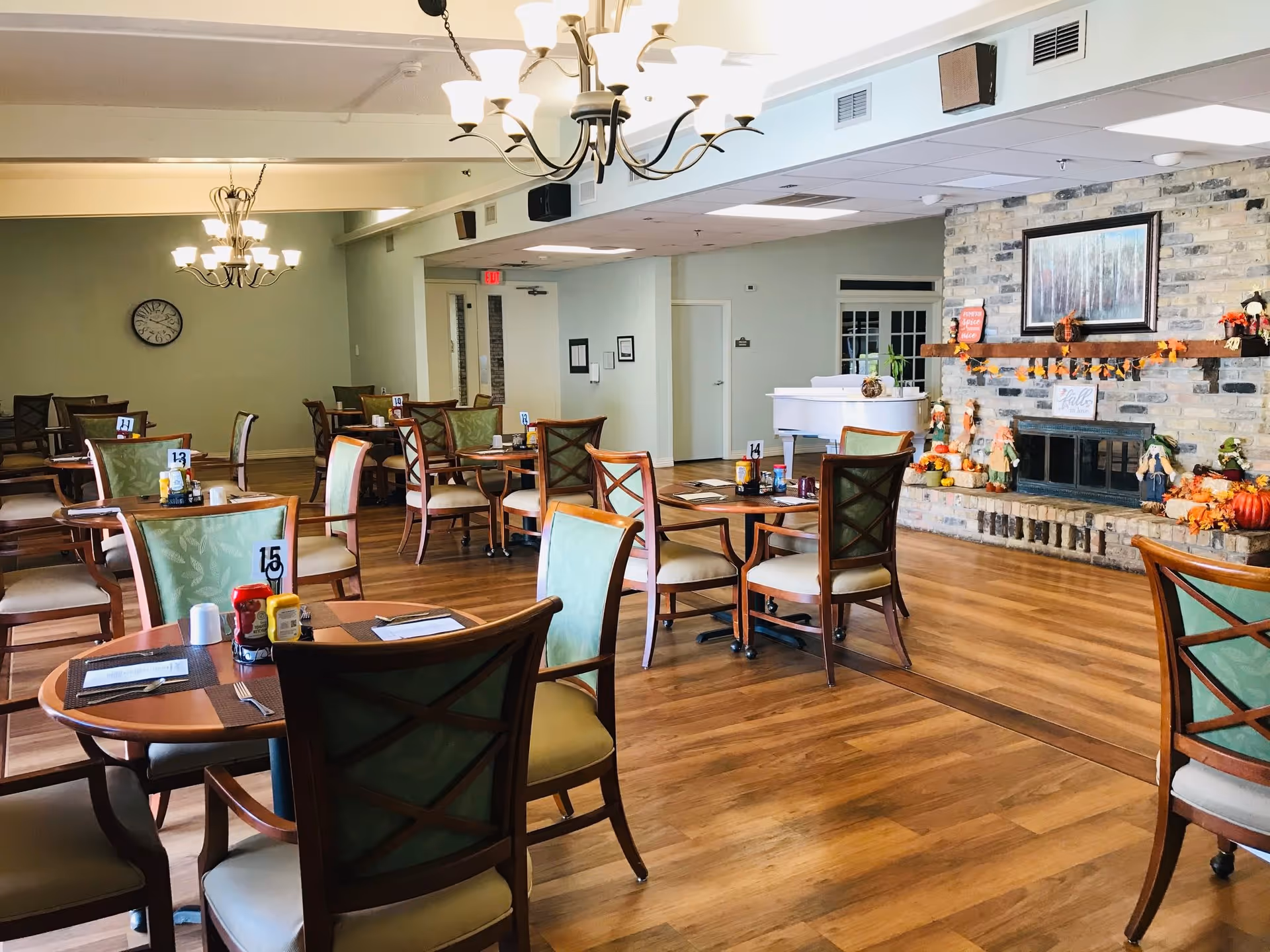 A dining room with multiple wooden tables and chairs arranged neatly on a wooden floor. Each table has placemats, menus, and condiments like ketchup and mustard. The room features two chandeliers hanging from the ceiling, a wall clock, and a brick fireplace decorated with autumn-themed ornaments and a framed painting above it. A white piano is positioned near the fireplace.