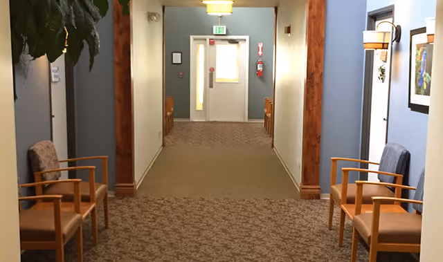 Carpeted hallway in an assisted living facility with chairs along the walls leading to a doorway with an exit sign.