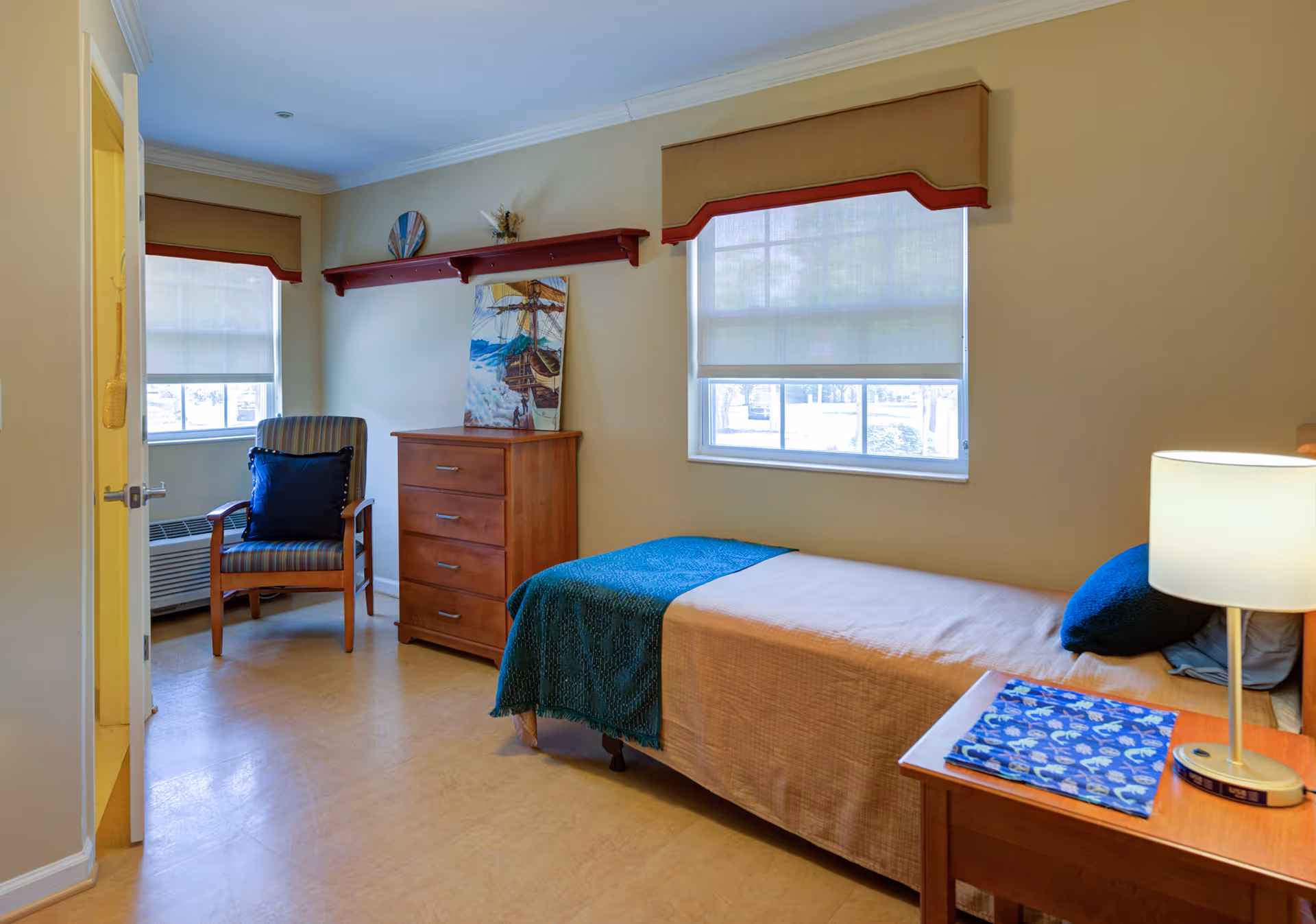 A small, cozy bedroom with a single bed covered with a beige bedspread and a teal blanket. There is a wooden nightstand with a lamp and a folded blue patterned cloth. A wooden dresser with four drawers is against the wall, with a painting and decorative items on top. A striped armchair with a dark blue cushion is placed near a window with beige and red valance curtains. The room has light-colored walls and flooring, with natural light coming through the windows.
