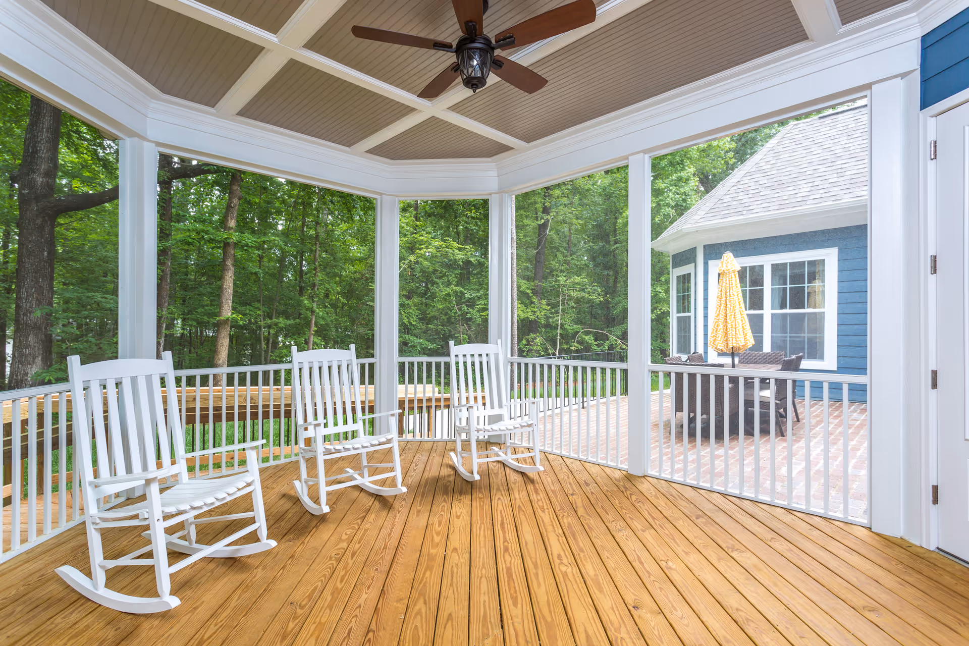 A screened-in wooden porch with three white rocking chairs and a ceiling fan. The porch overlooks a wooded area and an adjacent patio with a table, chairs, and a yellow umbrella next to a blue building.