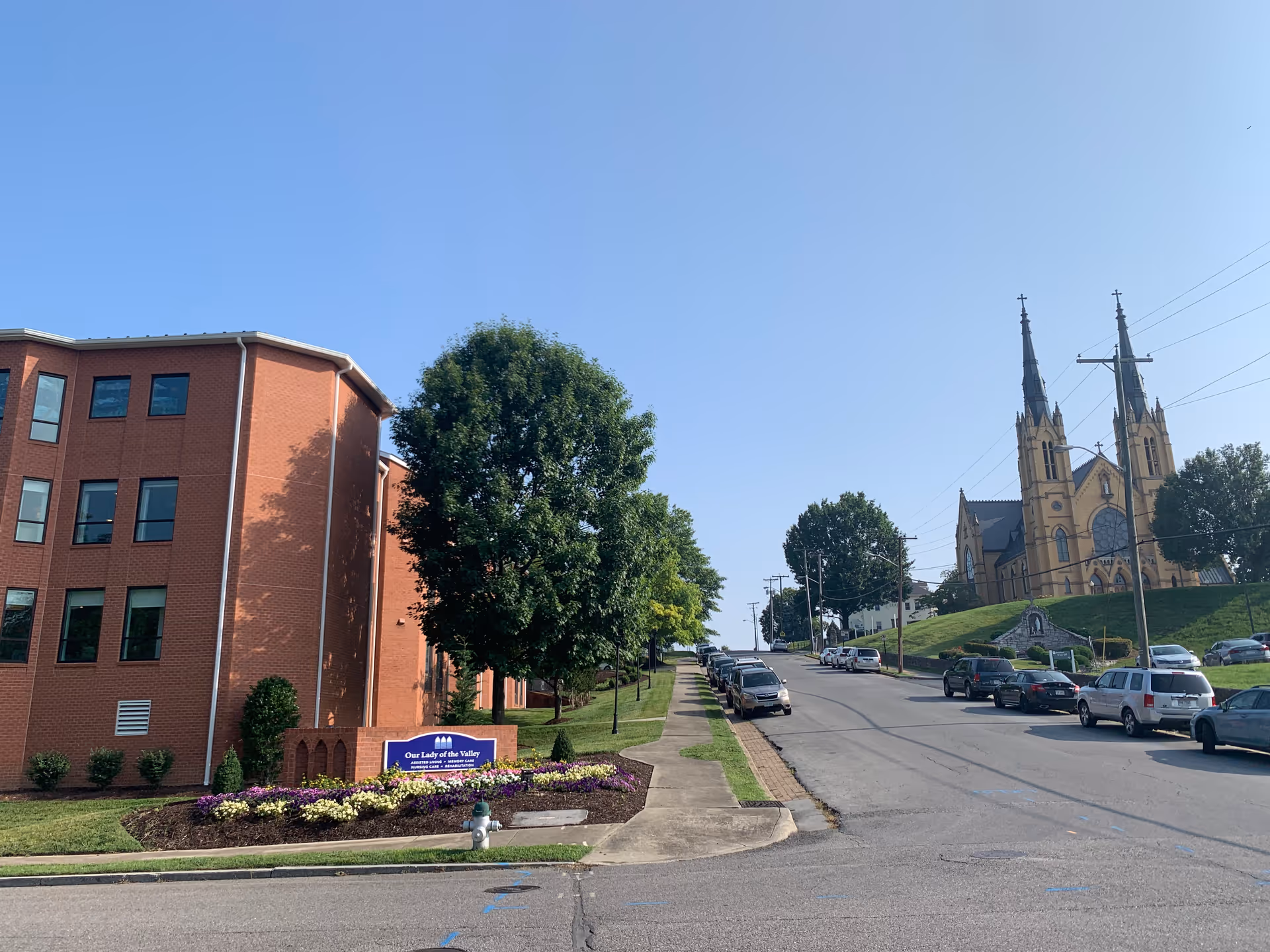 View of a street with a brick building on the left labeled Our Lady of the Valley, a landscaped flower bed in front, and a large church with two tall spires on a hill to the right. Several cars are parked along the street under a clear blue sky.