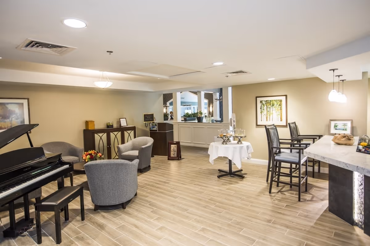 A spacious, well-lit common area in a senior living facility featuring a black grand piano with a bench, three gray upholstered armchairs arranged around a small table with a flower arrangement, a sideboard with decorative items, a round table covered with a white cloth holding glass cake stands, and a countertop with three high chairs. The walls are decorated with framed artwork, and the floor is covered with light wood-style tiles.