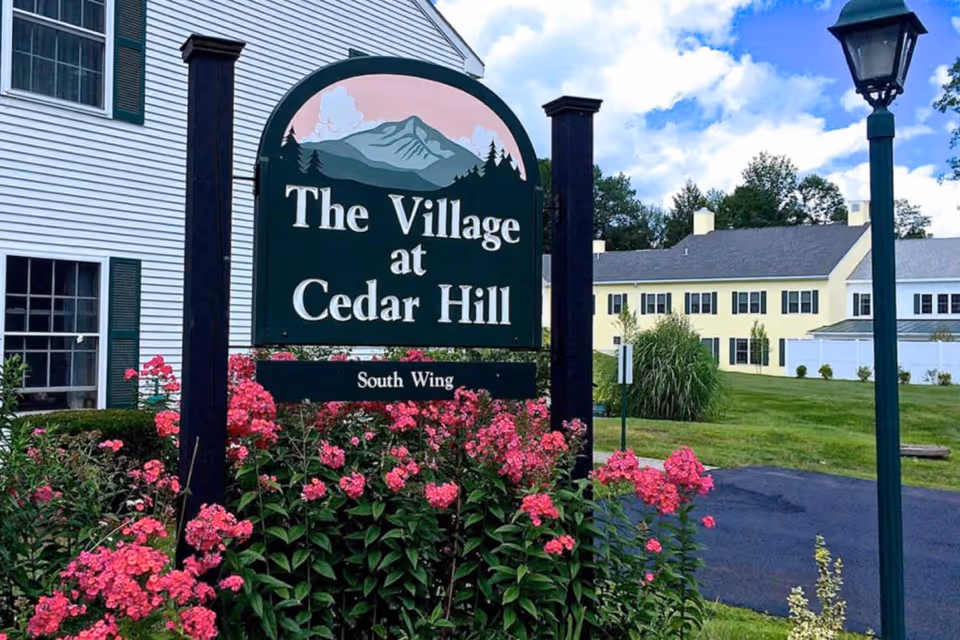 Entrance sign reading 'The Village at Cedar Hill' surrounded by pink flowers with the facility building and a lamp post in the background.