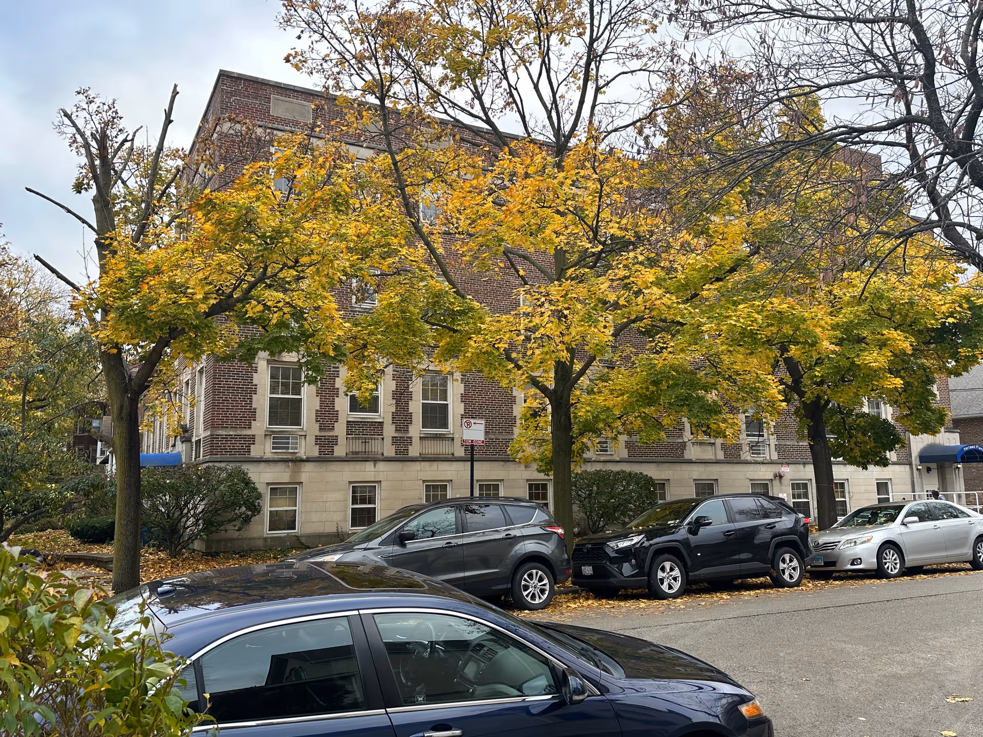Street view of a brick building partially obscured by trees with yellow autumn leaves. Several parked cars line the street in front of the building. The building has multiple windows and a blue awning near the entrance.