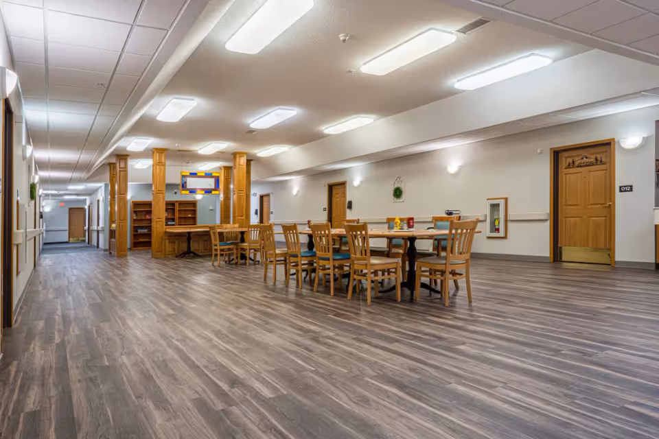 Spacious senior living common area with a long wood-look floor, wooden tables and chairs, and wood-paneled columns under bright ceiling lights.