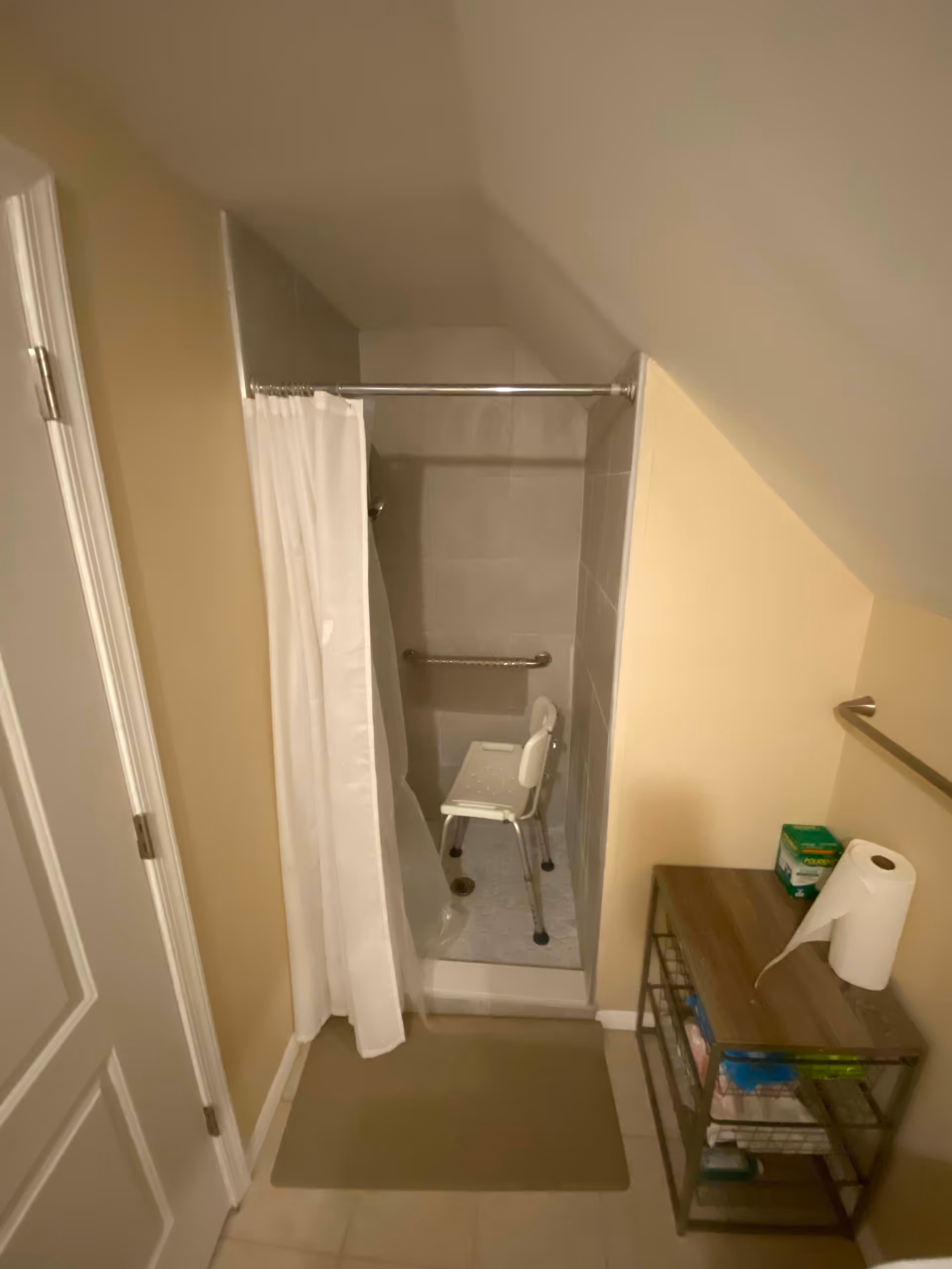 Small bathroom with a walk-in shower containing a white shower chair and grab bar, a partially closed shower curtain, floor mat, and a shelving unit with supplies.