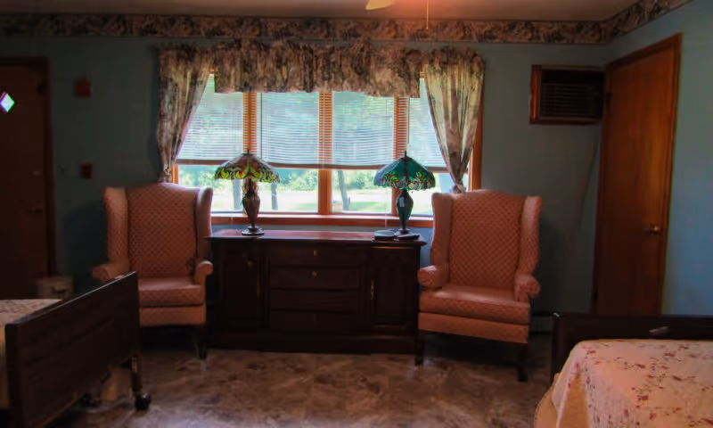 Bedroom with two pink wingback chairs flanking a wooden dresser beneath a window with floral curtains and lamps, with part of a bed visible.