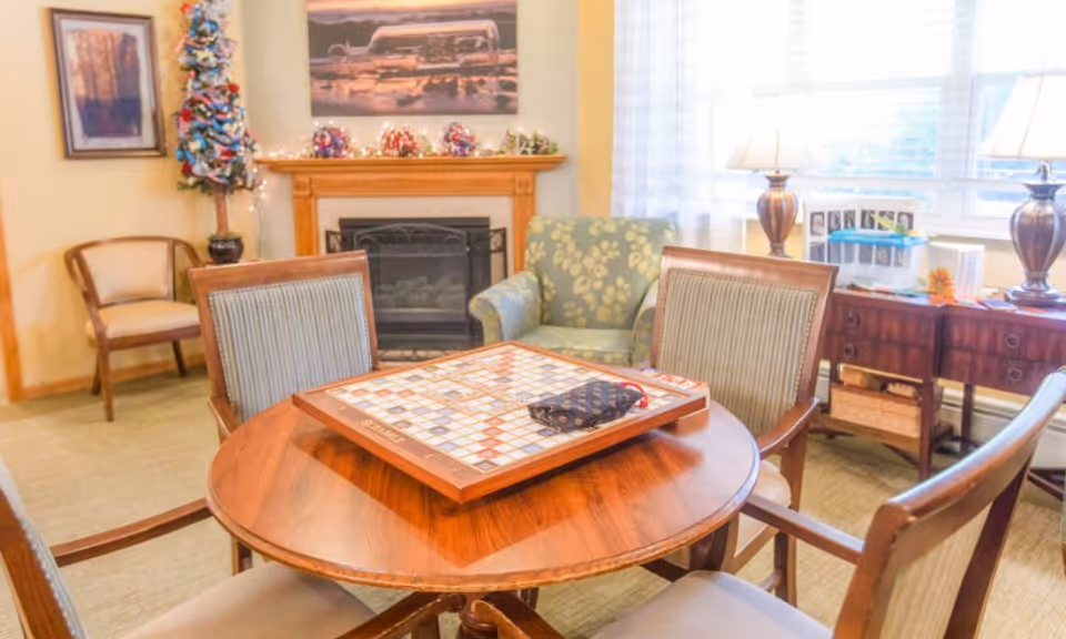A cozy living room area with a round wooden table in the center featuring a Scrabble board game. Four wooden chairs with cushions surround the table. In the background, there is a fireplace with decorative items on the mantel, a green armchair with a floral pattern, a small decorated Christmas tree, framed artwork on the walls, and a wooden sideboard with two lamps and various items on top. Light filters in through large windows with sheer white curtains.