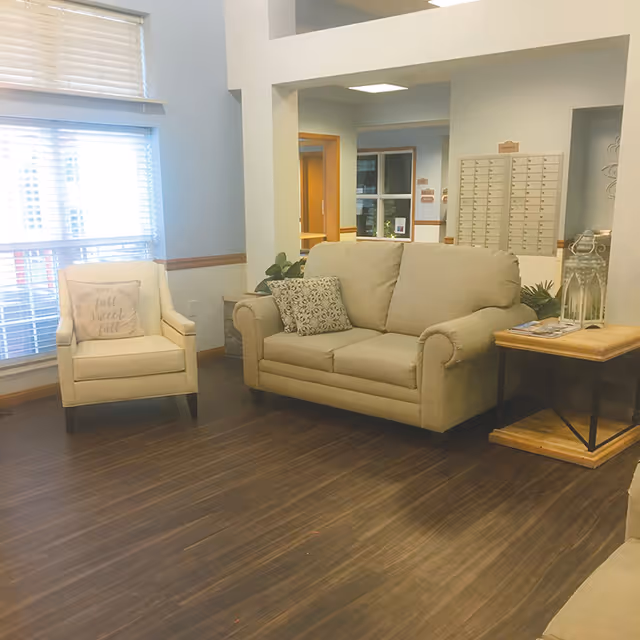 A cozy seating area in a senior living facility with a beige loveseat and an armchair. The armchair has a decorative pillow with the text 'fall sweet fall'. There is a wooden side table with a lantern and some magazines. The room has wooden flooring, light blue walls, and a window with blinds allowing natural light to enter.