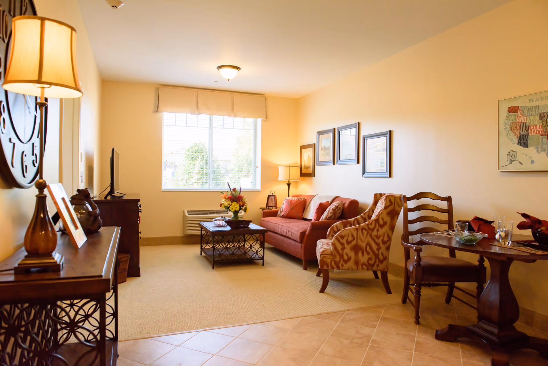 A cozy living room in a senior living facility with a red sofa, patterned armchair, wooden coffee table with a flower arrangement, and a small dining table with two chairs. The room has beige walls, a large window with blinds, framed artwork on the walls, and a floor lamp providing warm lighting.